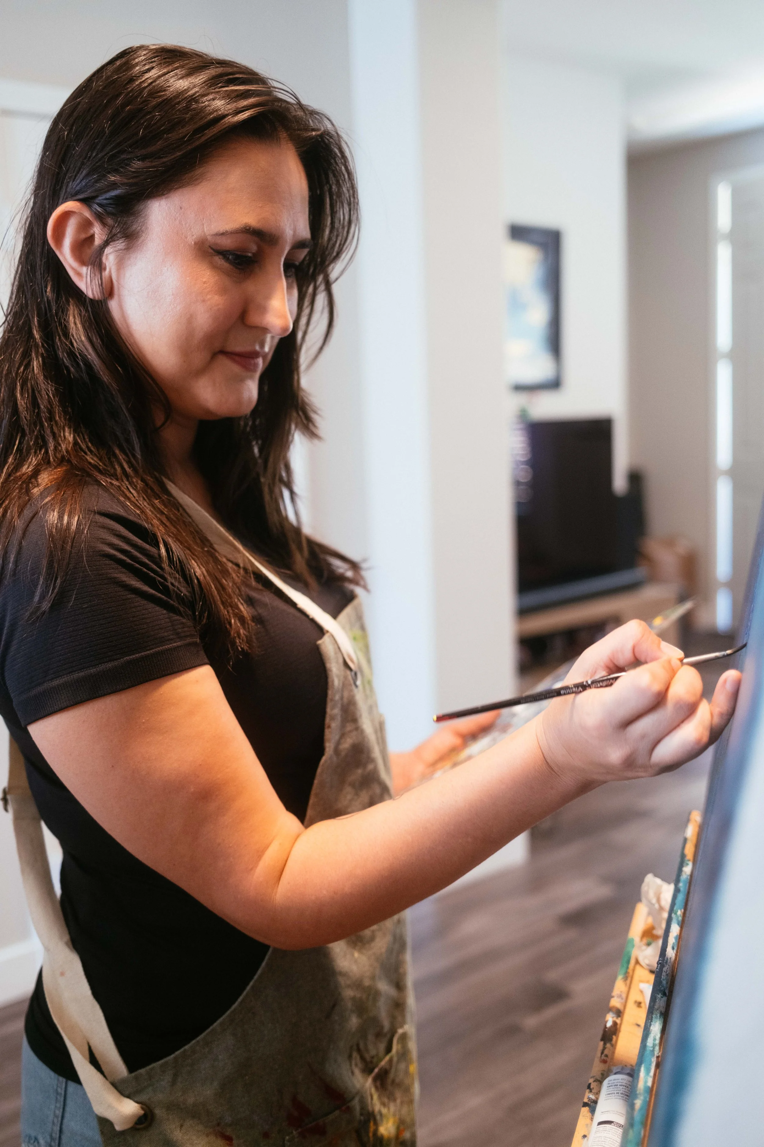 A woman with brown hair wearing an apron and black shirt standing in a room, painting on a canvas with a paintbrush.