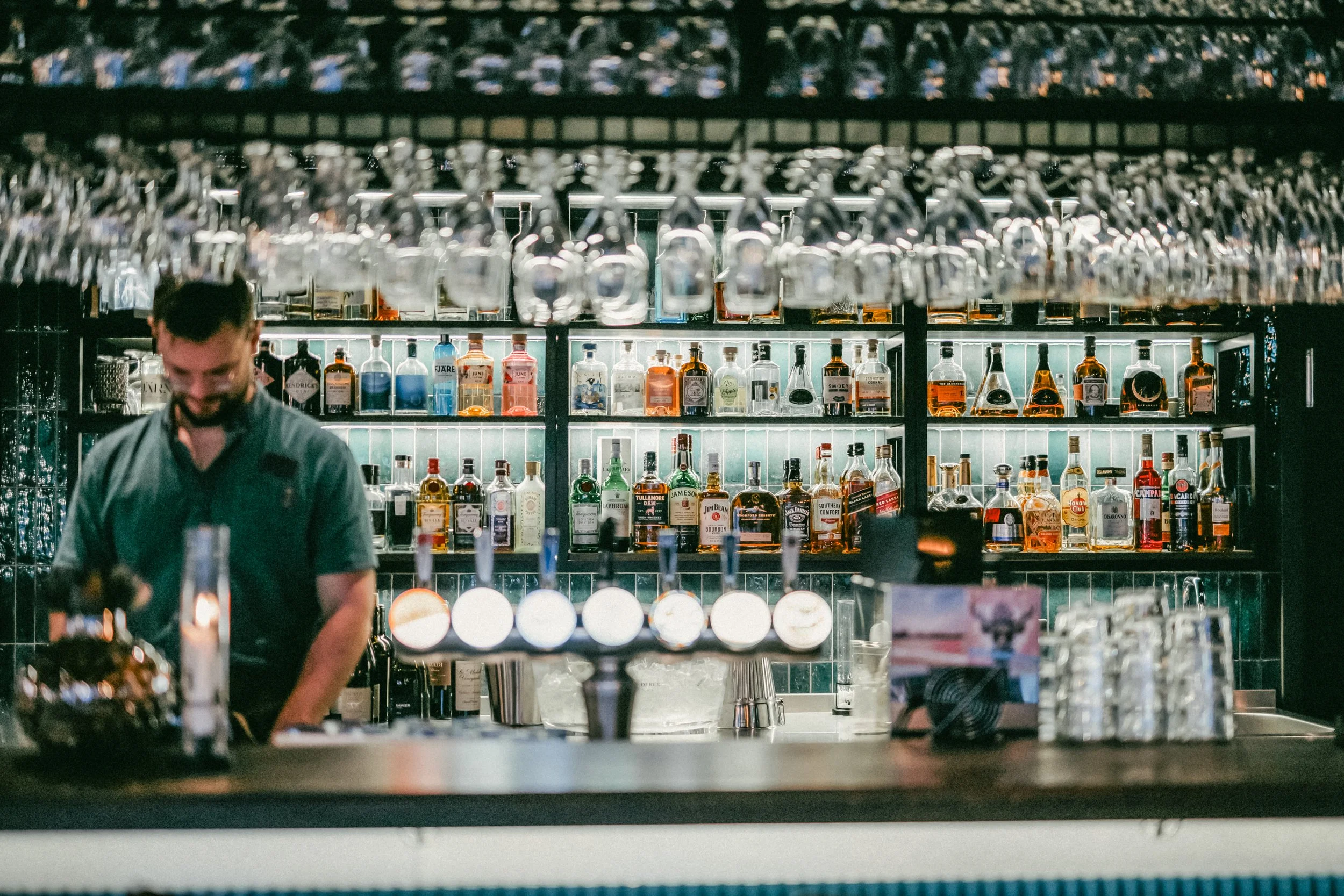 En bartender står bakom en bar med en bakgrund av hyllor fyllda med olika sorters sprit och glas, samt en öppen bar med olika flaskor och dekorationer.
