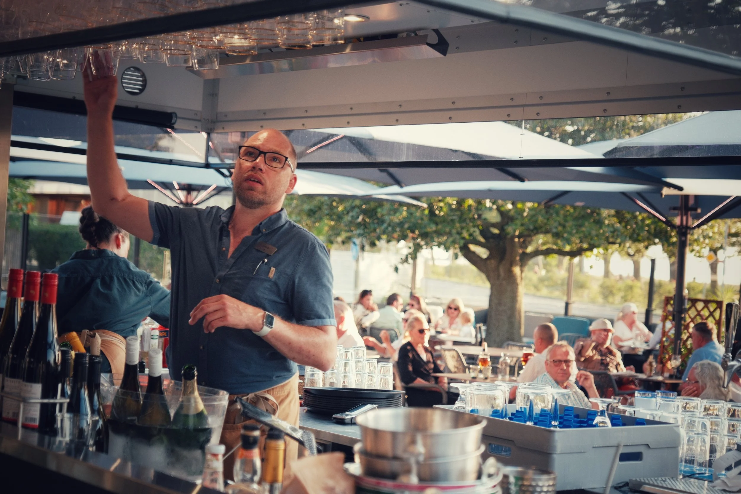 Man som arbetar bakom ett serveringsställe utomhus med folk sittande i bakgrunden, träd och skärmar