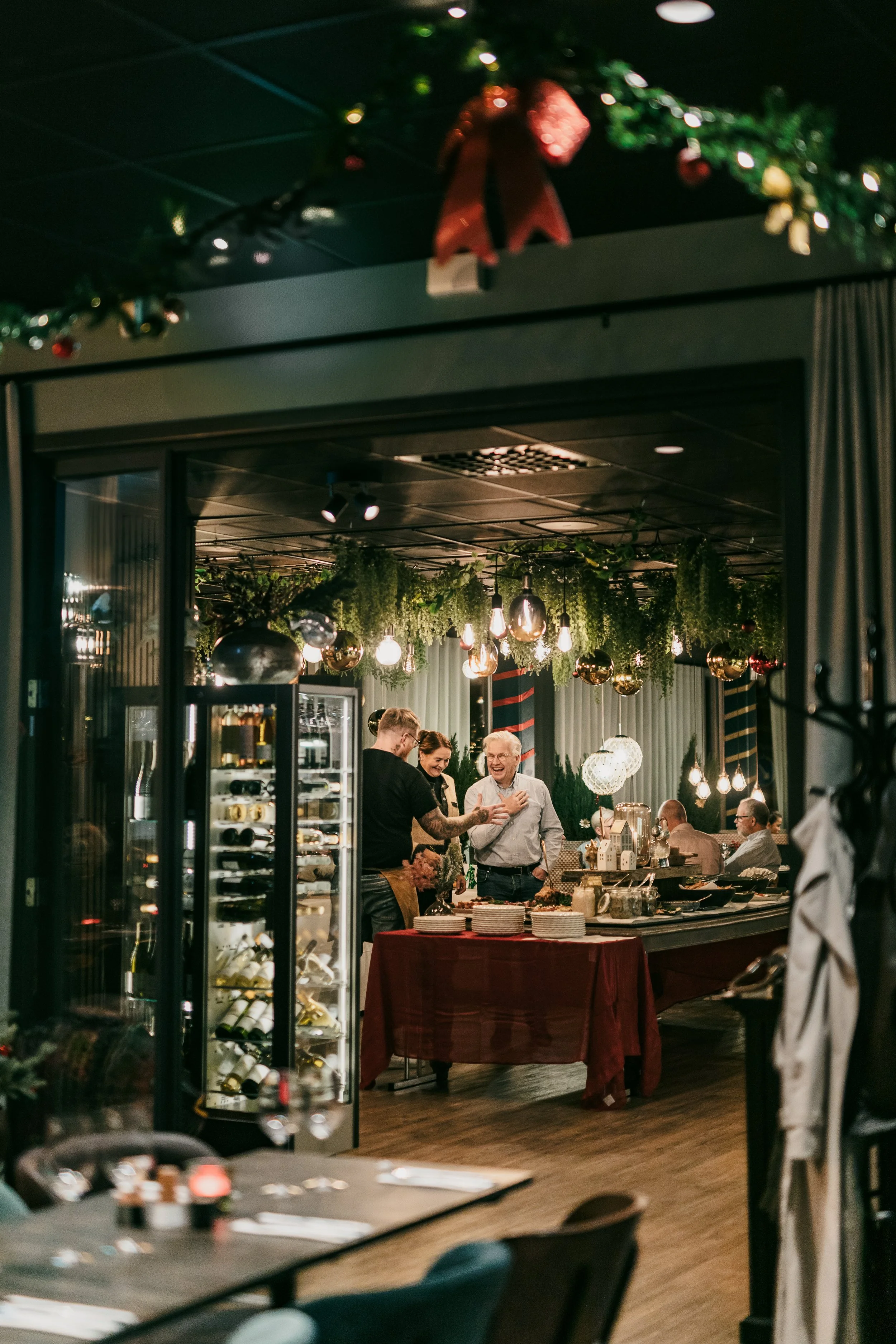 Människor som firar jul i ett festligt restauranginteriör, dekorerat med grönt och julstjärnor, med flera personer som skrattar och pratar vid ett buffébord.