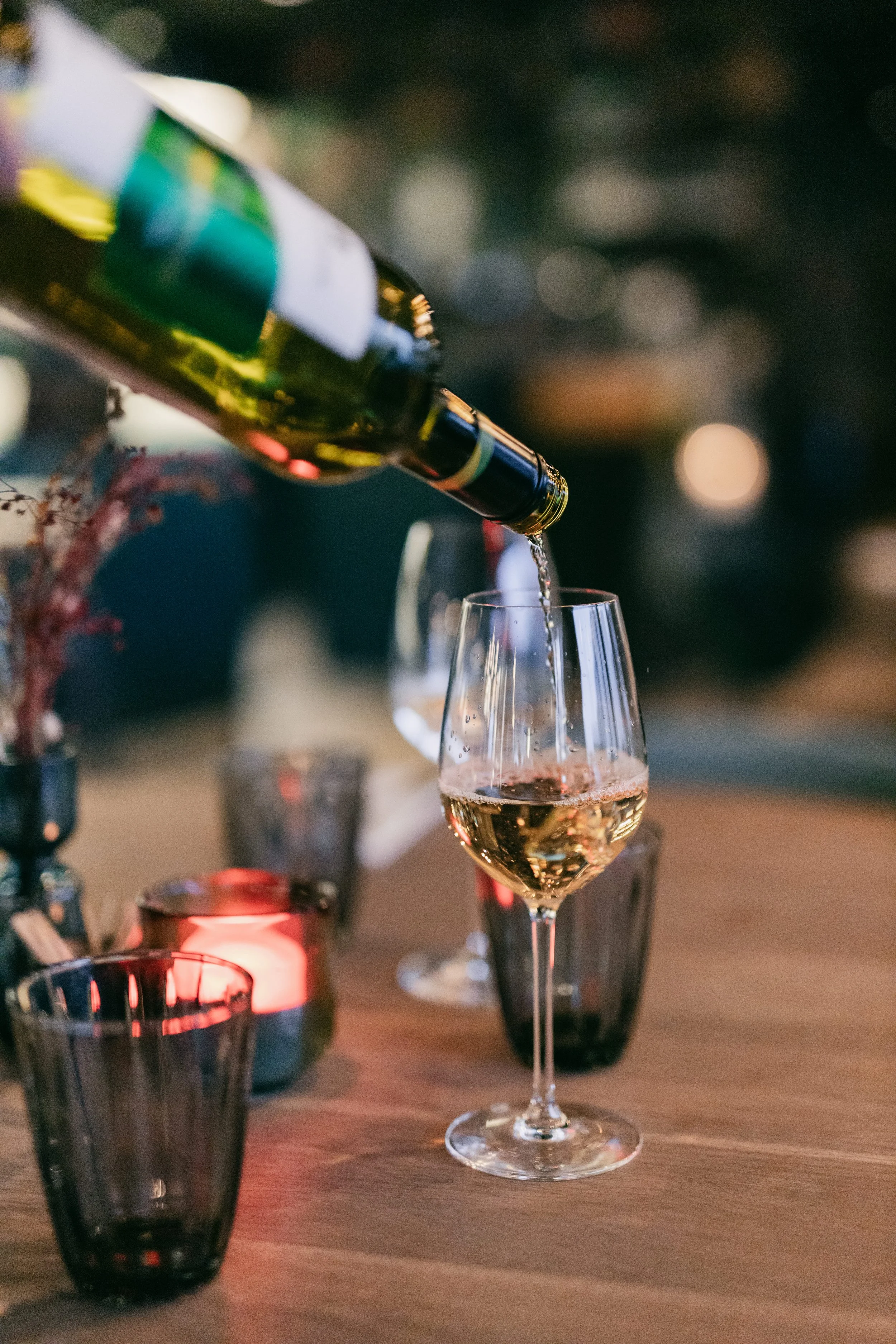 En flaska vitt vin som hälls i ett vinglas på ett middagsbord, omgivet av ytterligare glas och levande ljus.