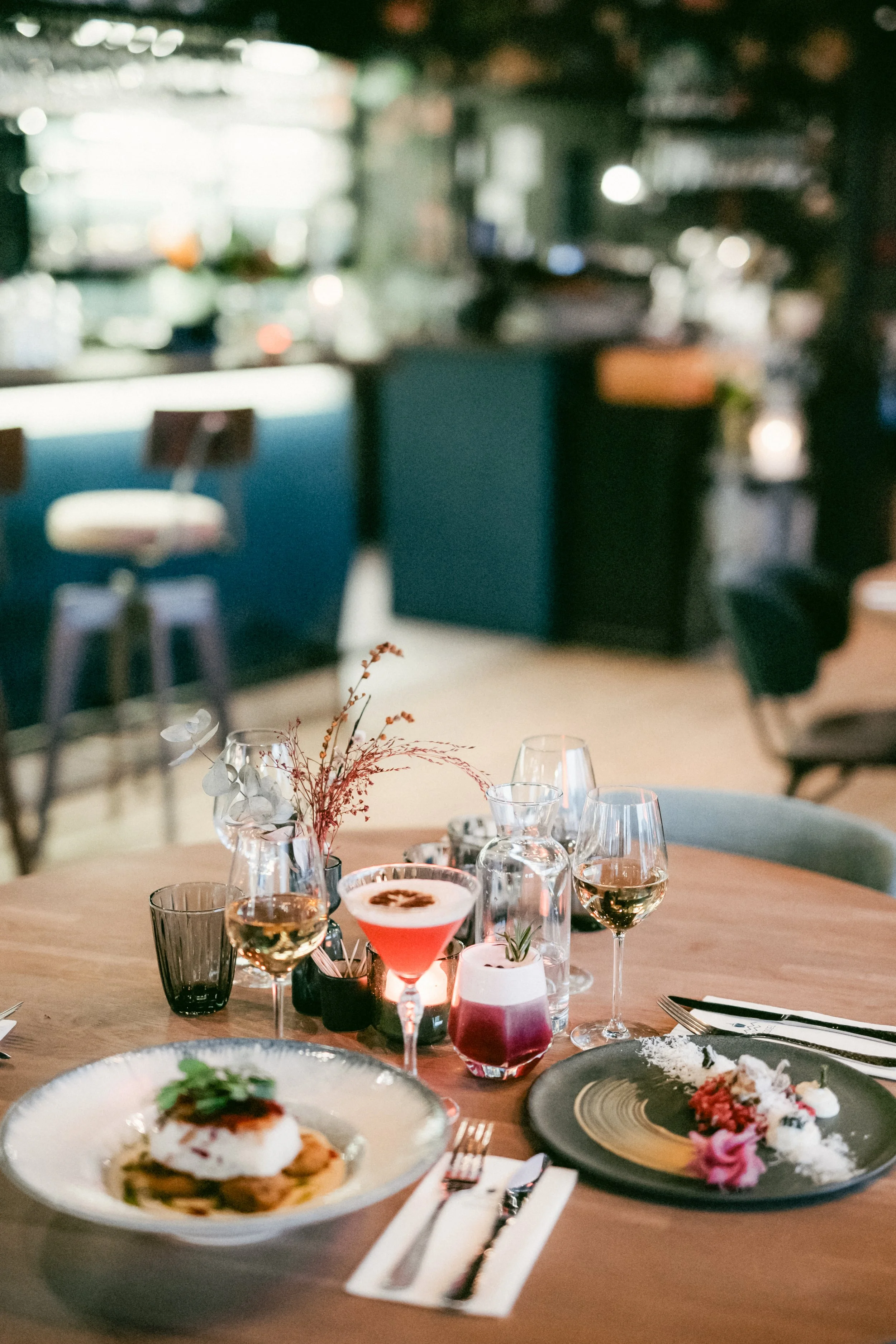 Elegant middagsbord uppdukat med glas, vaser med blommor, en cocktail, en vinflaska, och en tallrik med mat, i en restaurangmiljö.