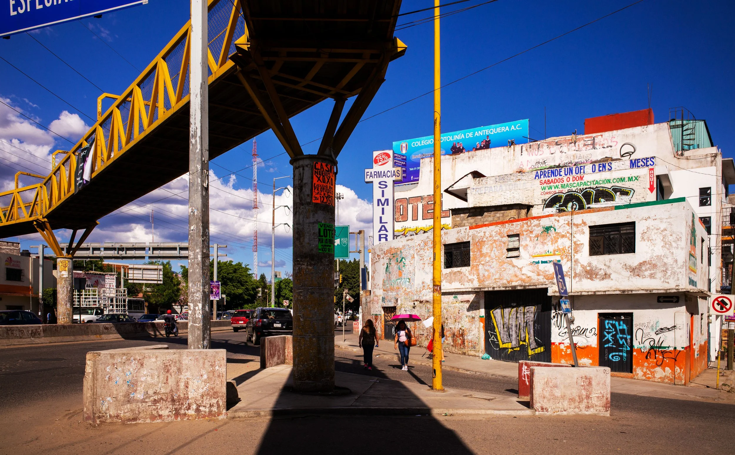 City street scene with an elevated yellow pedestrian bridge, people walking, cars on the road, and colorful signs on buildings against a bright blue sky.