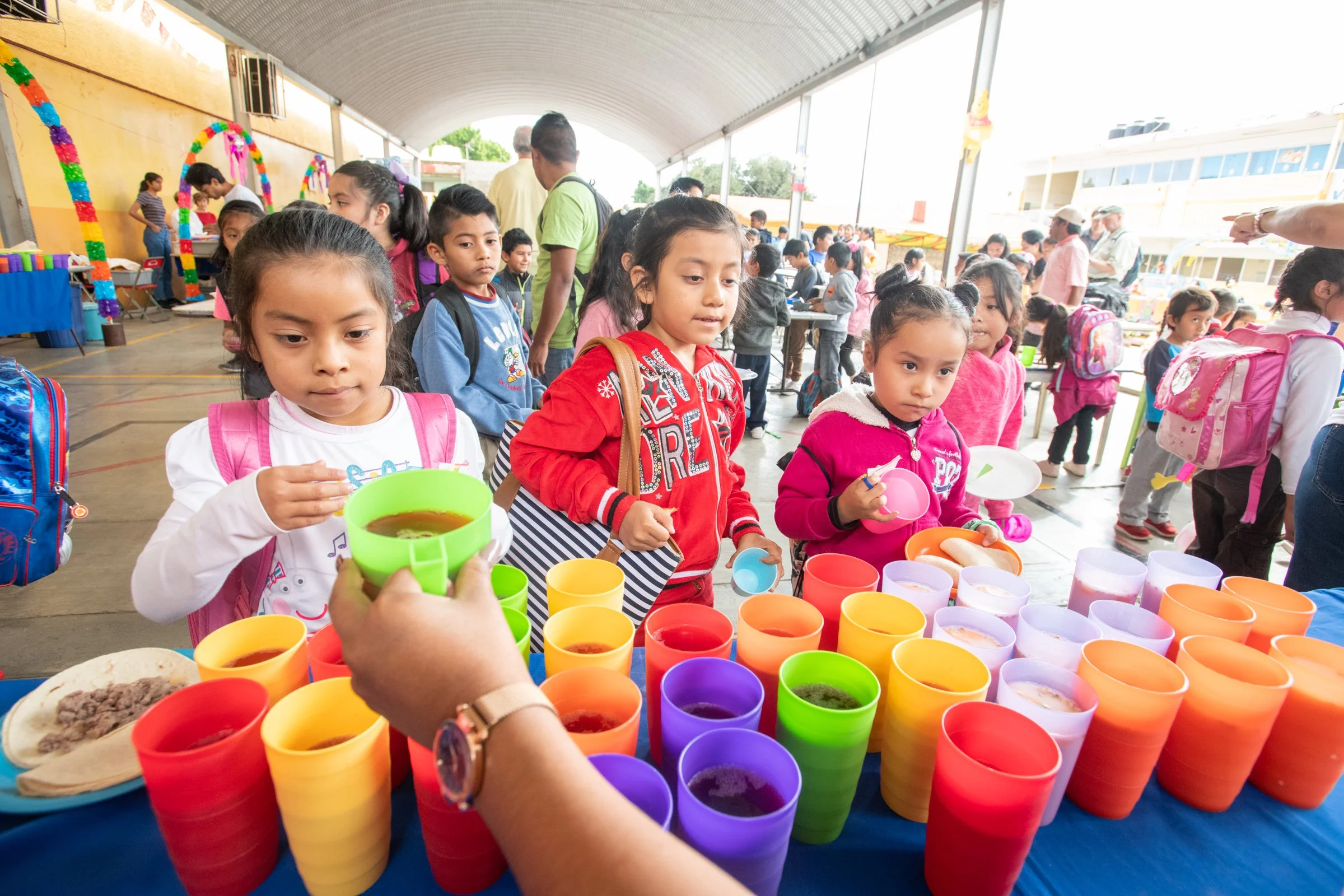 Children at a school event are lining up to receive colorful cups with a drink under a covered outdoor area.