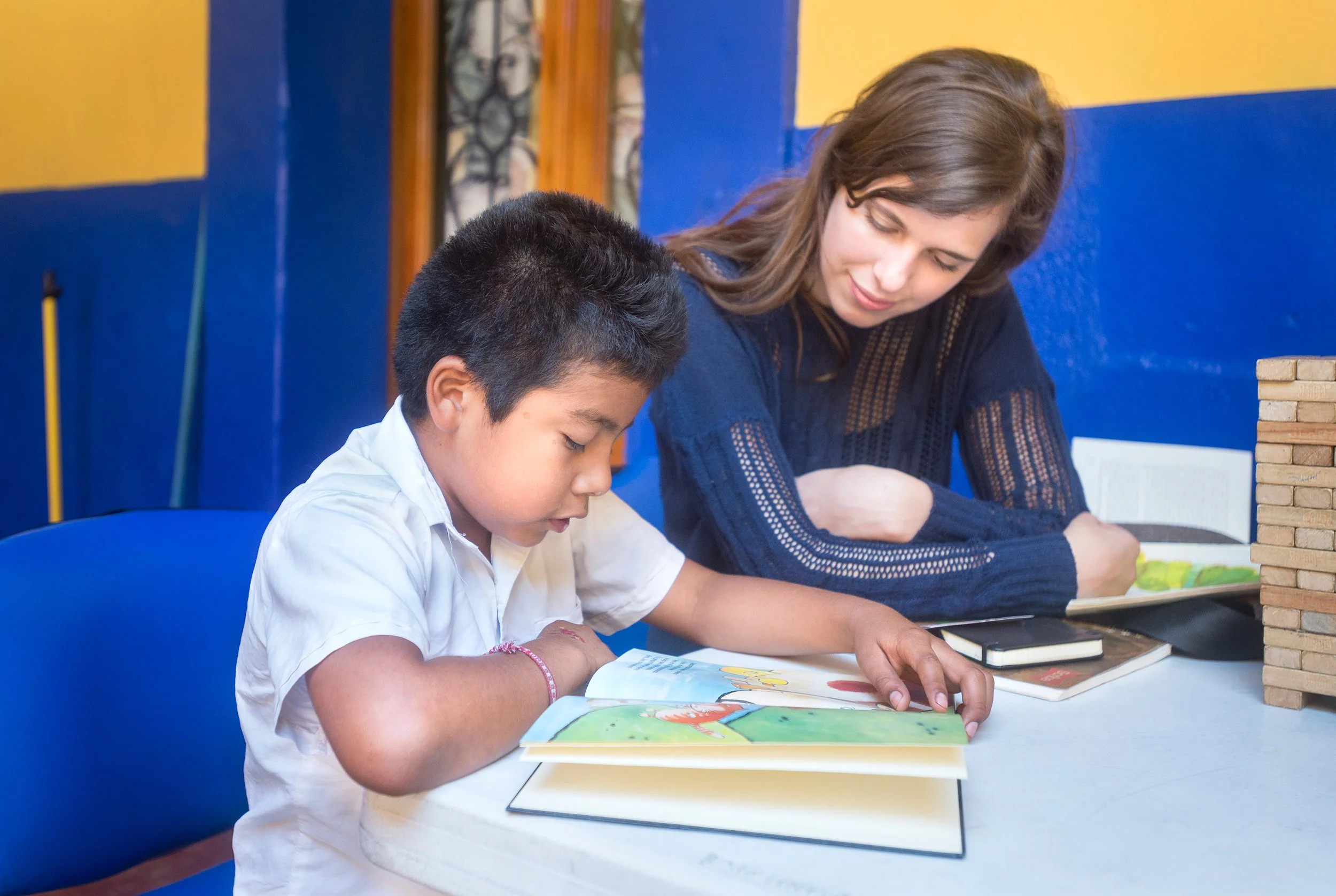A teacher and a student are sitting at a table, reading a book together in a classroom at Casa Jodi.