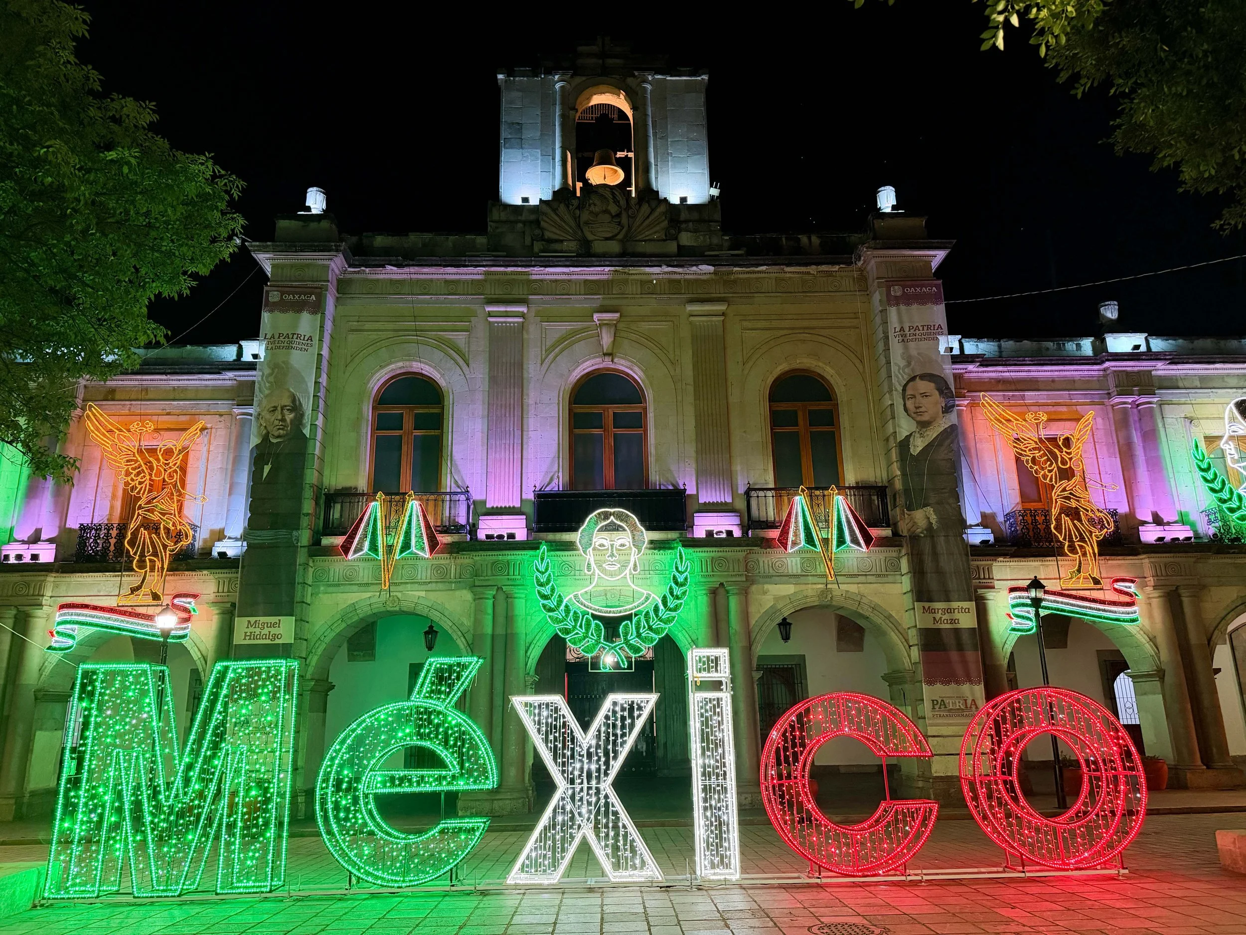A large illuminated Mexican flag in green, white and red, featuring the national coat of arms, displayed outdoors at night.