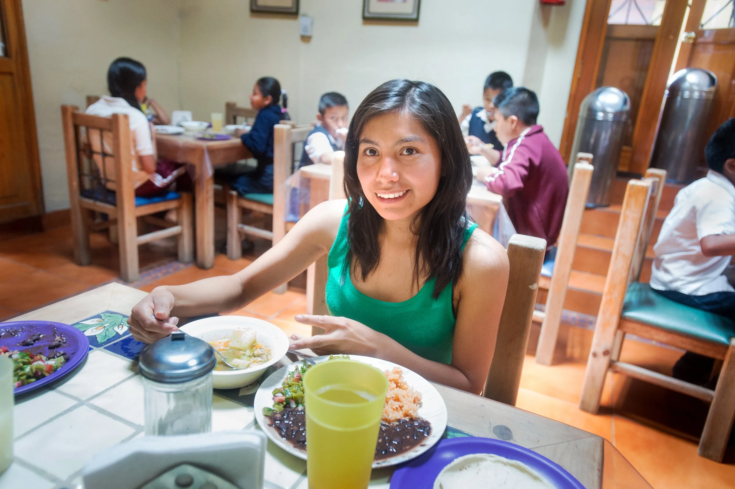A girl is sitting at a cafeteria table with plates of food at Centro de Esperanza Infantil (Center of Hope for Children, or CEI) with children dining in the background.