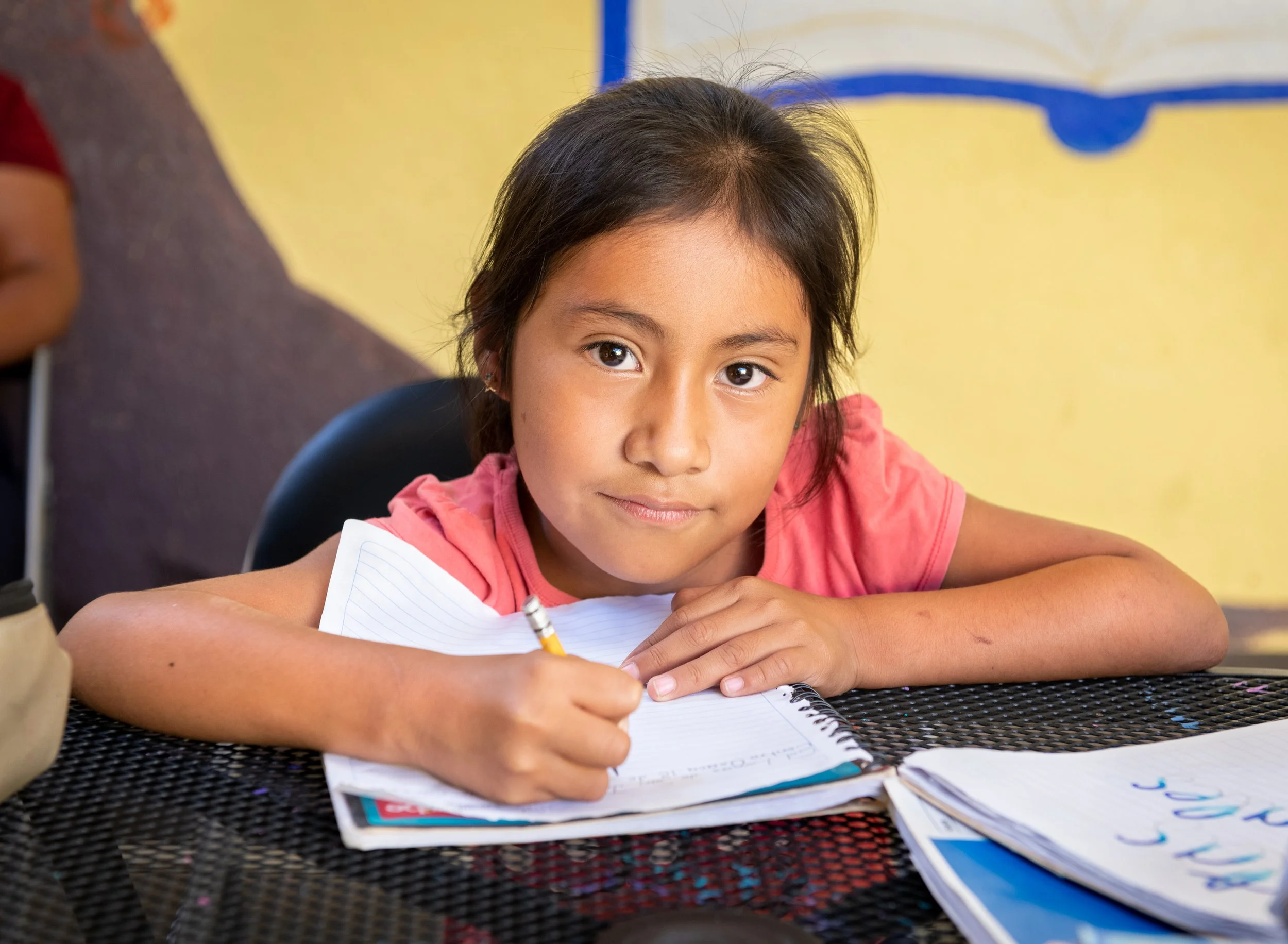 A young girl is sitting at a table at Casa Jodi, holding a pencil and writing in a notebook.