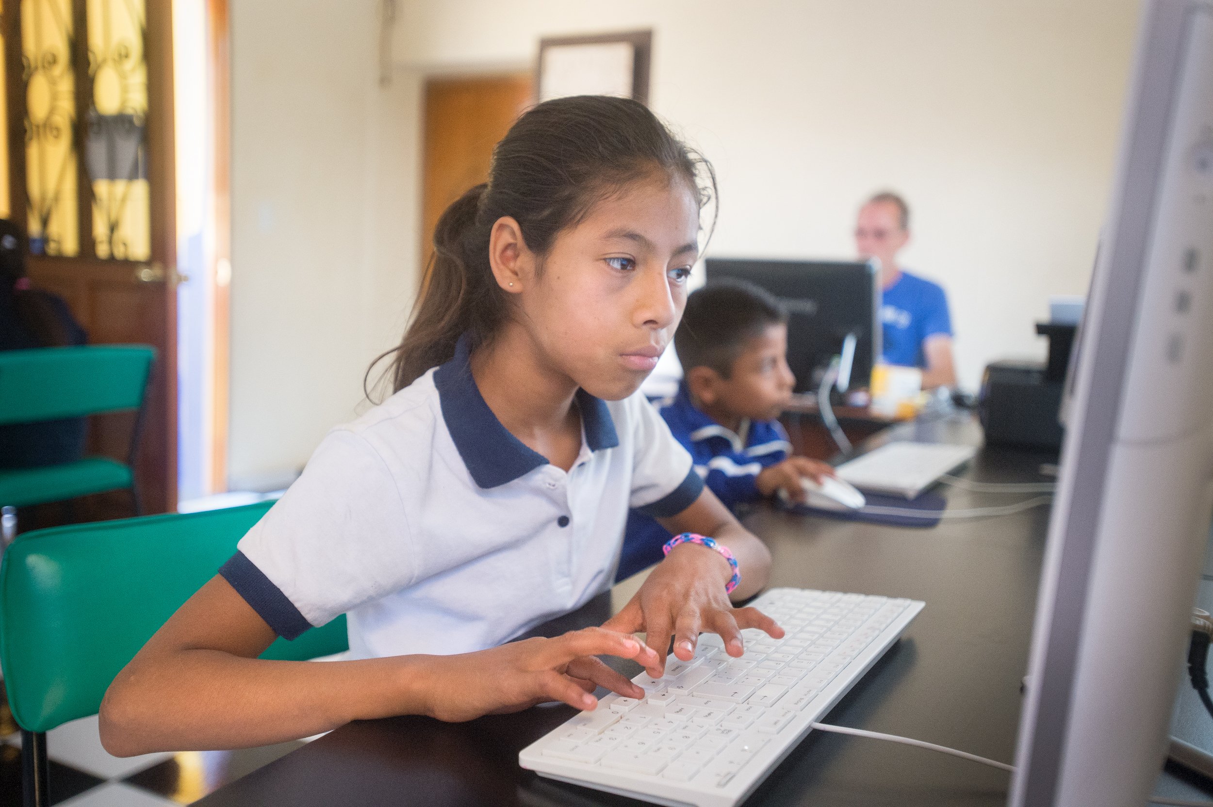A young girl is working on a computer in a classroom at Casa Jodi with other students and an instructor in the background.