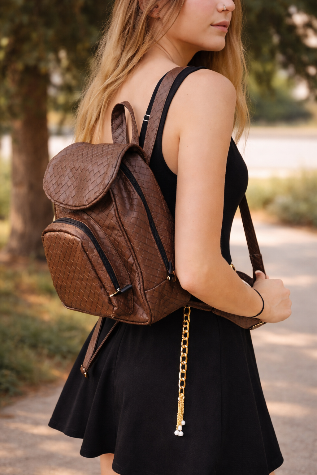 A woman with blonde hair and a nose ring, wearing a black sleeveless dress, carries a brown woven backpack with a flap and zipper pockets, along with a gold chain accessory hanging from her waist, outdoors with trees and a pathway in the background.