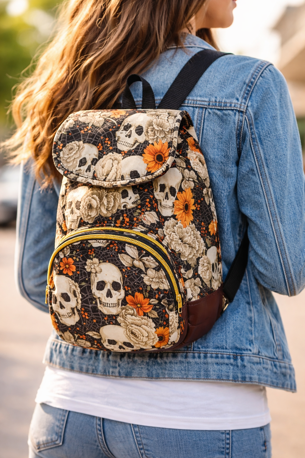 A woman with long wavy brown hair wearing a denim jacket and jeans, carrying a backpack with skull, flower, and web patterns, outdoors in daylight.