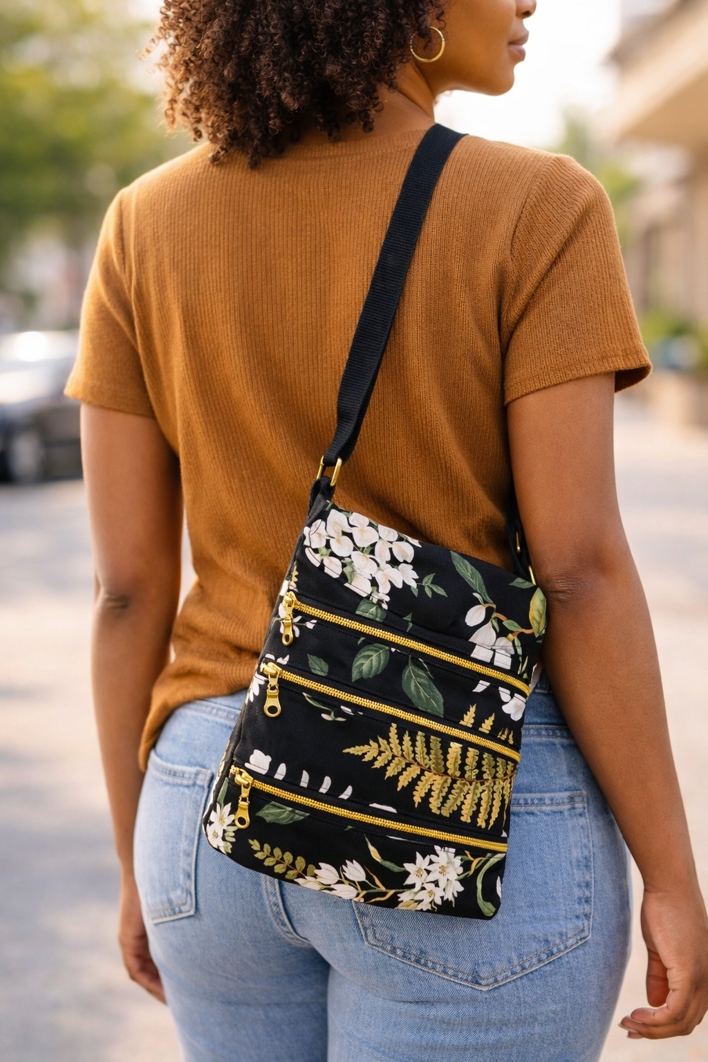 A woman wearing a brown short-sleeve top and light blue jeans with a black floral crossbody bag with gold zippers.