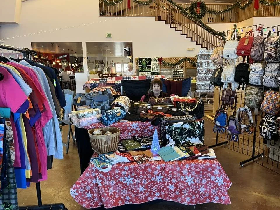A Christmas-themed craft fair booth with handbags and accessories, decorated with holiday items, set inside a store with hanging ornaments and a staircase with holiday garland.