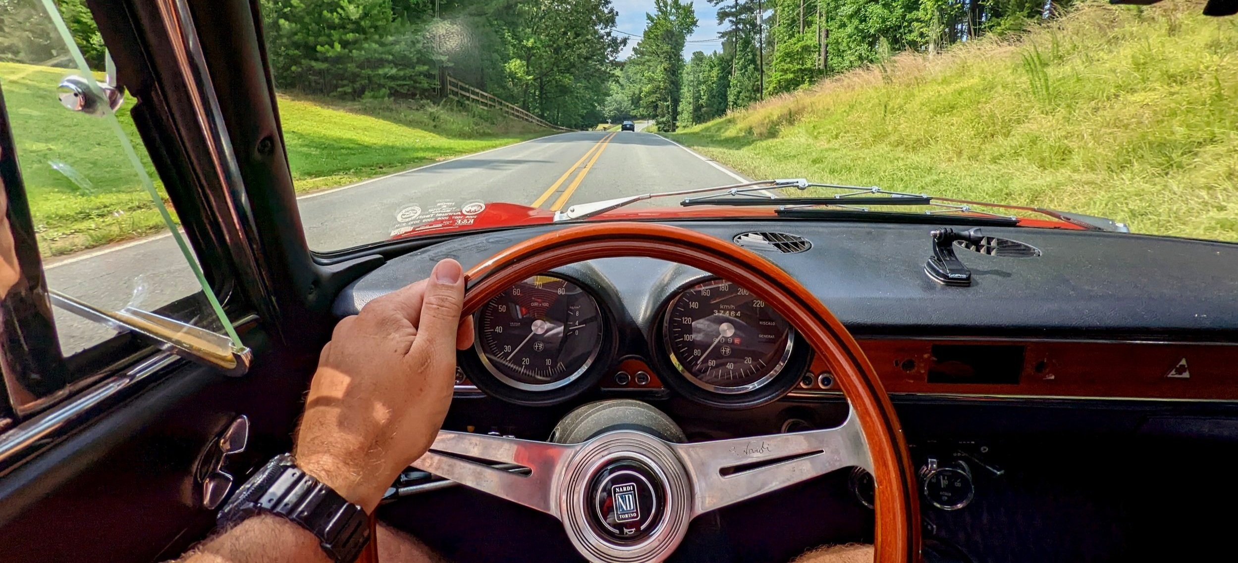 Interior view from a vintage car showing the dashboard, steering wheel, and driver’s hand on the wheel, on a sunny road surrounded by green trees and grass.