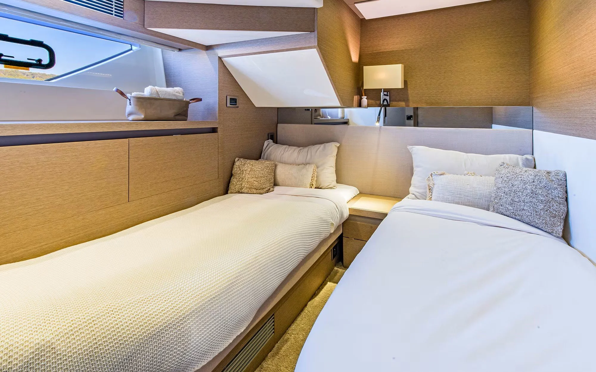 Two beds in a modern hotel room with beige headboards, pillows, a nightstand, window with a basket of towels, and wall lamps.
