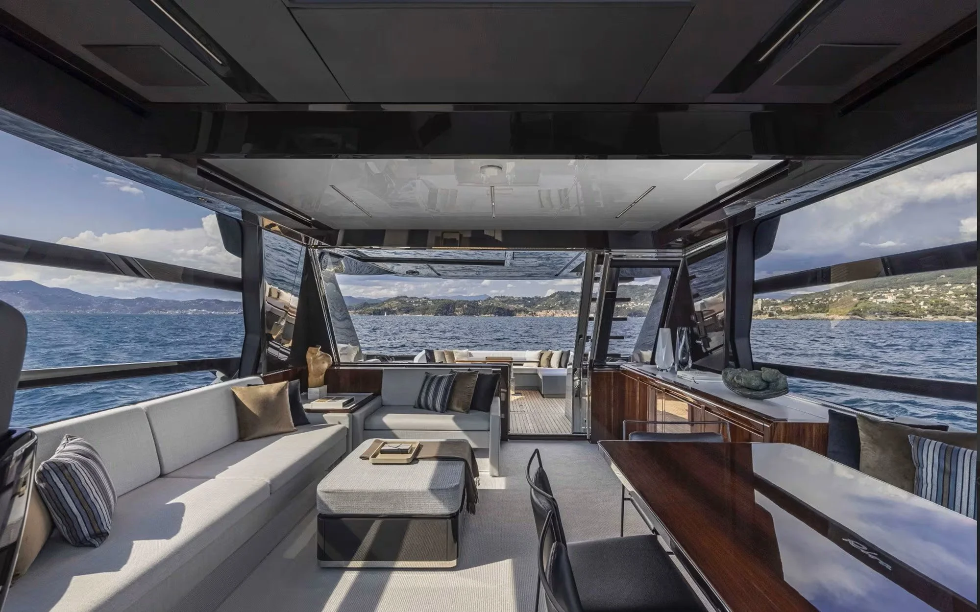 Luxury yacht interior with large windows showing ocean and distant shoreline, featuring modern furnishings including a white sofa with pillows, a wooden table with a marble top, and a small ottoman.