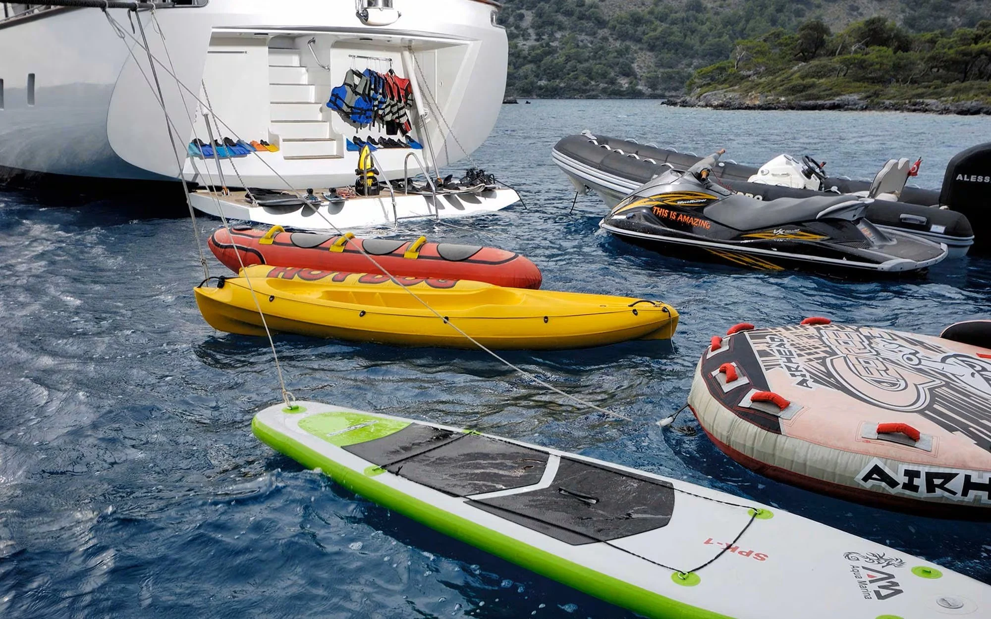 Various watercrafts including a large yacht, kayaks, jet skis, and inflatable boats floating on the water near a rocky shoreline.