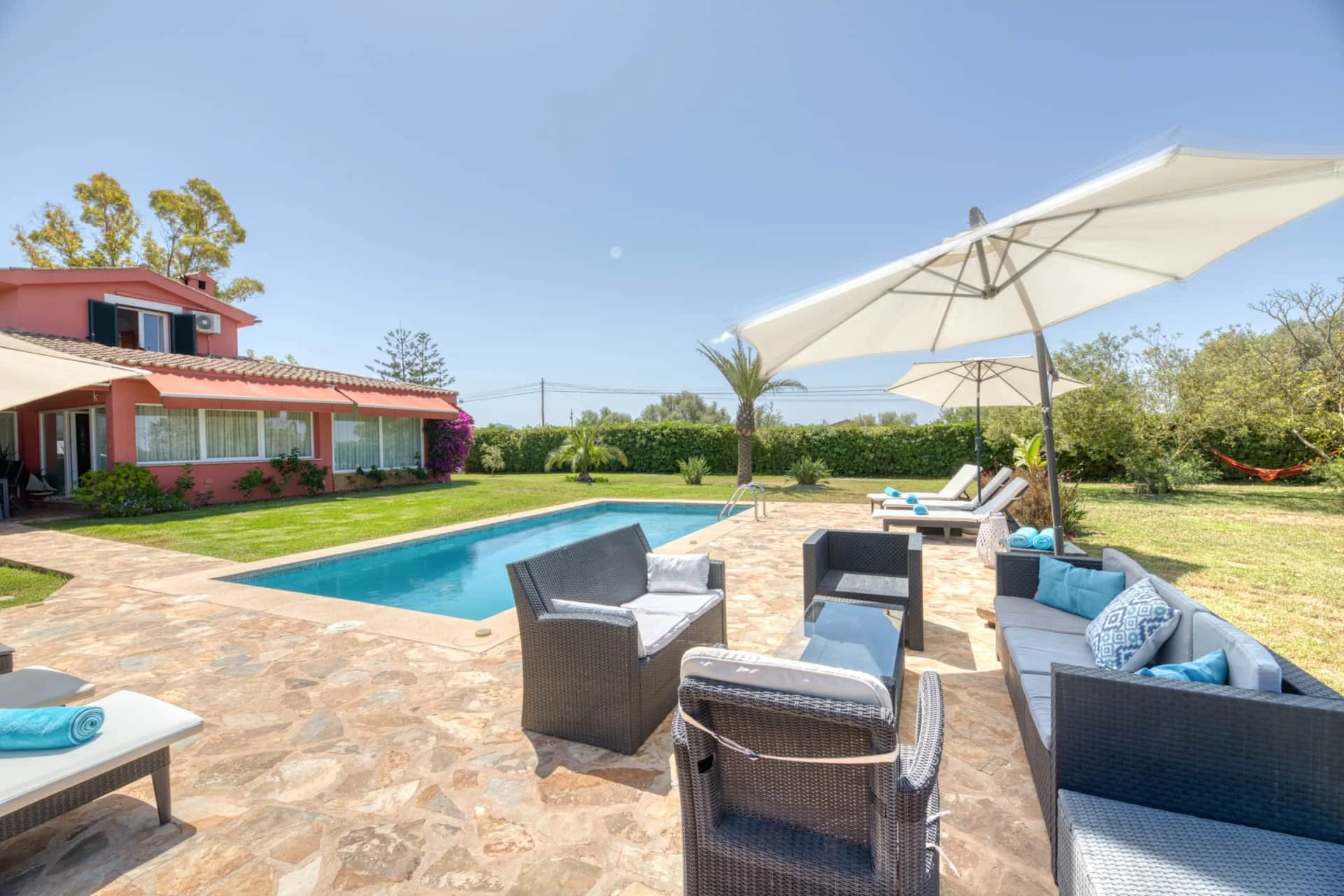 A backyard with a swimming pool, outdoor furniture, large umbrellas, lush green grass, palm trees, and a pink house in the background under a clear blue sky.