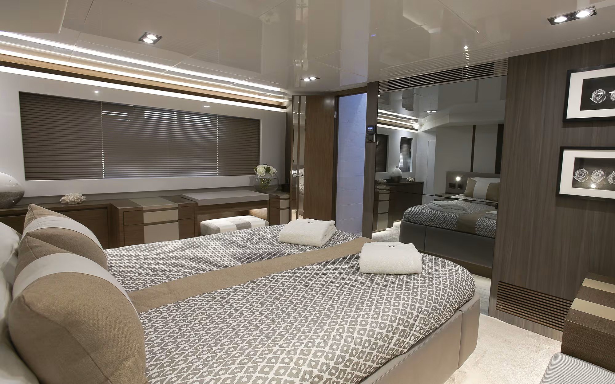 A luxurious hotel bedroom featuring two neatly made beds with patterned bedding, plush pillows, and towels. The room has modern decor with artwork on the wall, a window with blinds, and a mirrored wall element adding to the upscale ambiance.