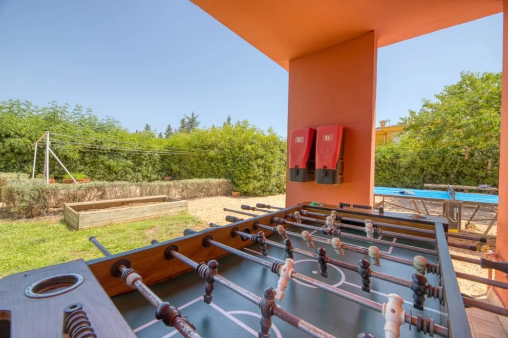 Foosball table outdoors on patio with greenery, garden bed, and pool in background during daytime.