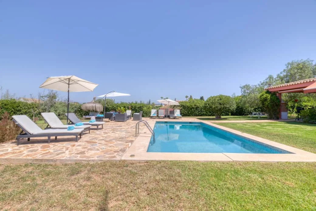 Outdoor swimming pool area with lounge chairs, umbrellas, and a grassy lawn under a clear blue sky.