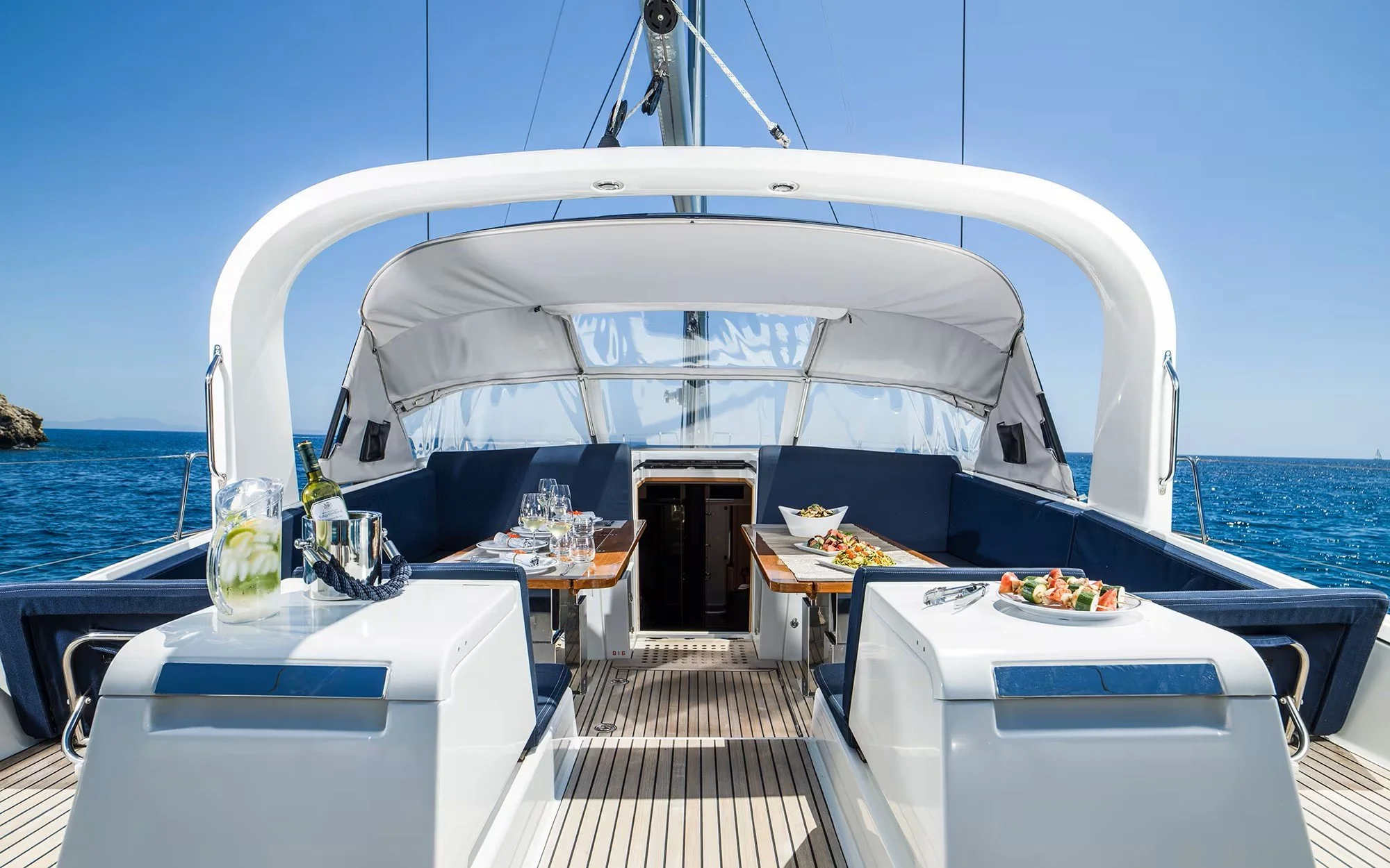 On a yacht, a dining area with tables set with wine glasses, plates of food, and a pitcher of beverage, with a view of the ocean and a clear blue sky.