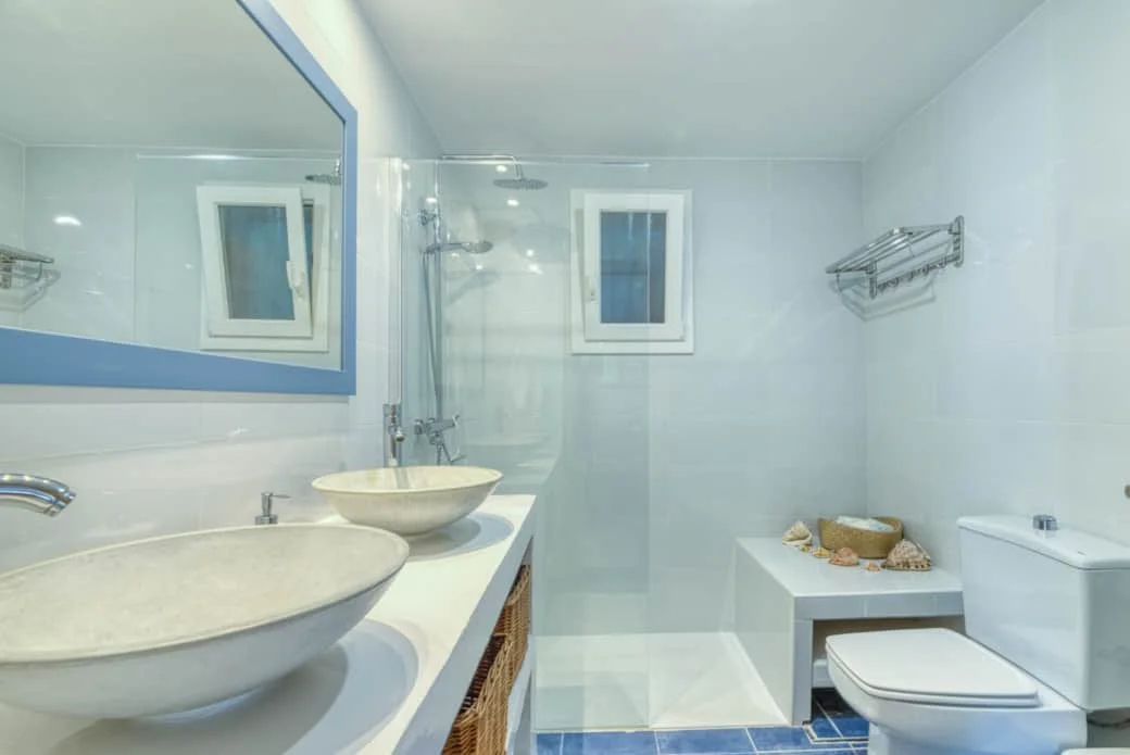 Modern bathroom with two vessel sinks, a large mirror, a walk-in shower with a small window, a toilet, and decorative shells on a bench.