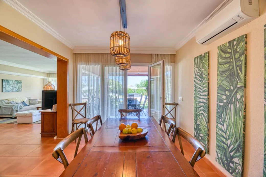 Dining room with a wooden table and six chairs, hanging pendant lights, sliding glass door leading to a balcony, and tropical leaf artwork on the right wall.