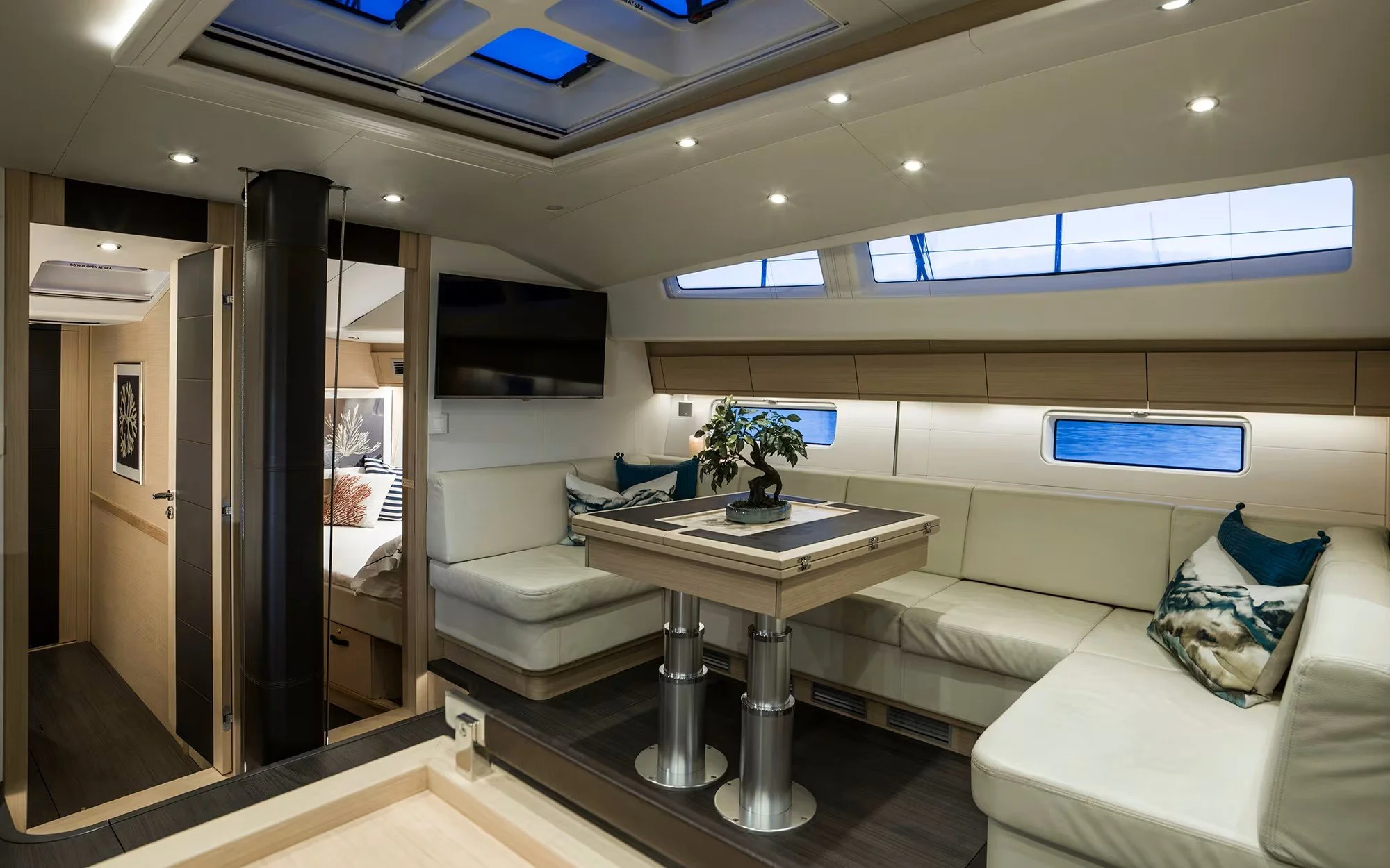 Interior of a yacht with a white L-shaped sofa, a flat-screen TV on the wall, a small table with a bonsai plant, and windows revealing water outside. There is a bedroom visible through an open doorway.