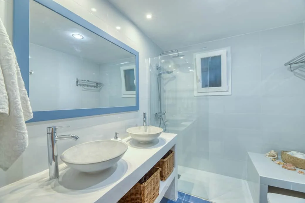 Bright, modern bathroom with two vessel sinks, large mirror with a blue frame, and a shower area with a glass divider and small window.