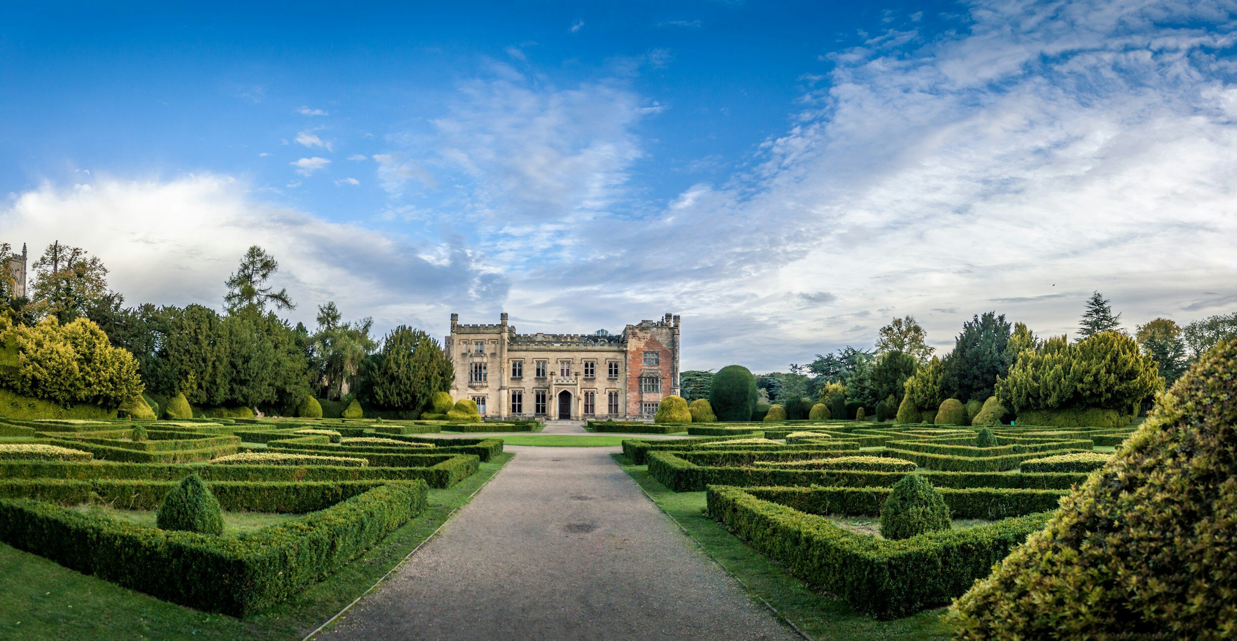 A historic castle surrounded by a beautifully maintained formal garden with neatly trimmed hedges and lush green trees, under a partly cloudy sky.