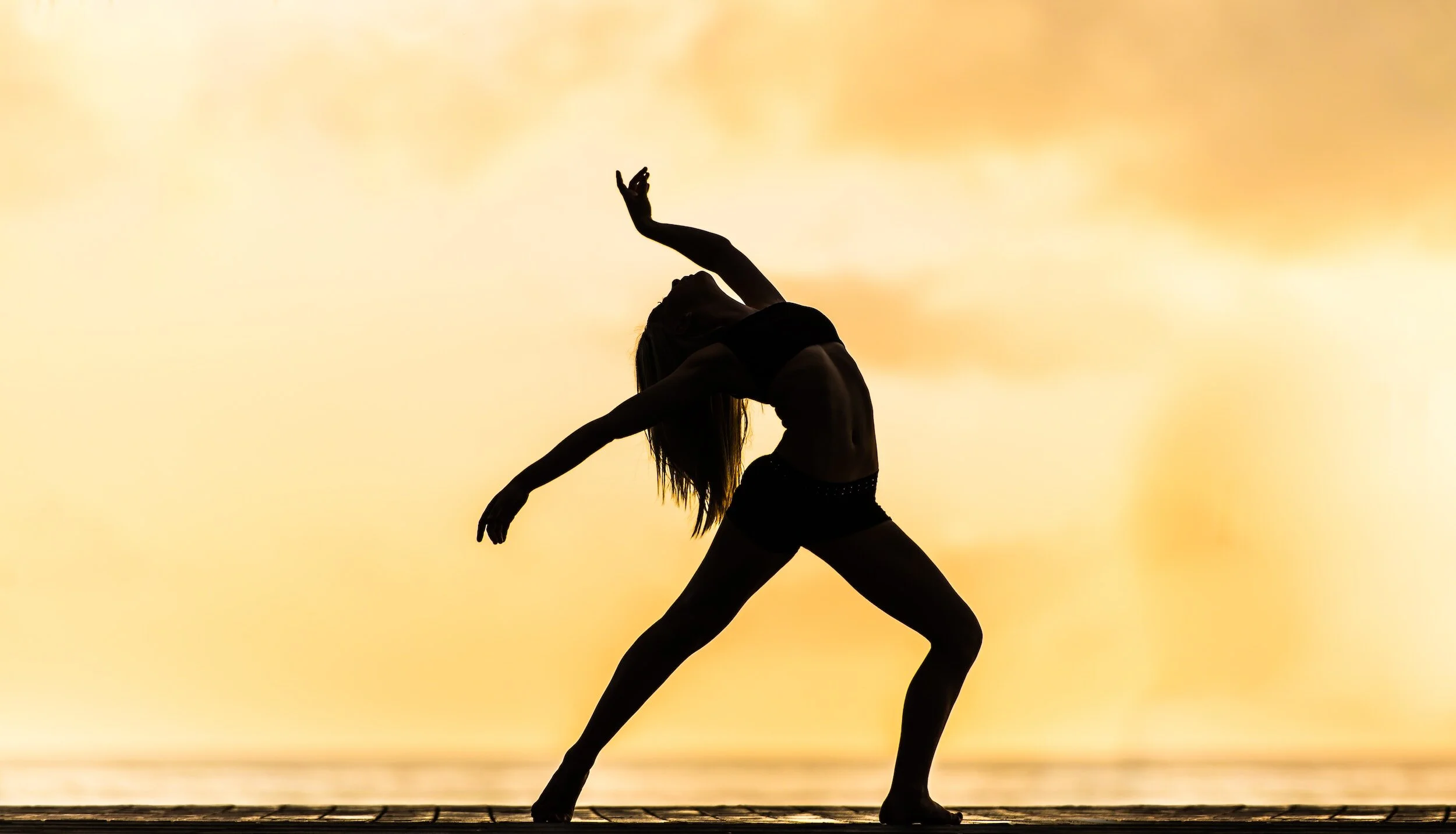 Silhouette of a woman dancing or practicing yoga at sunset by a body of water.