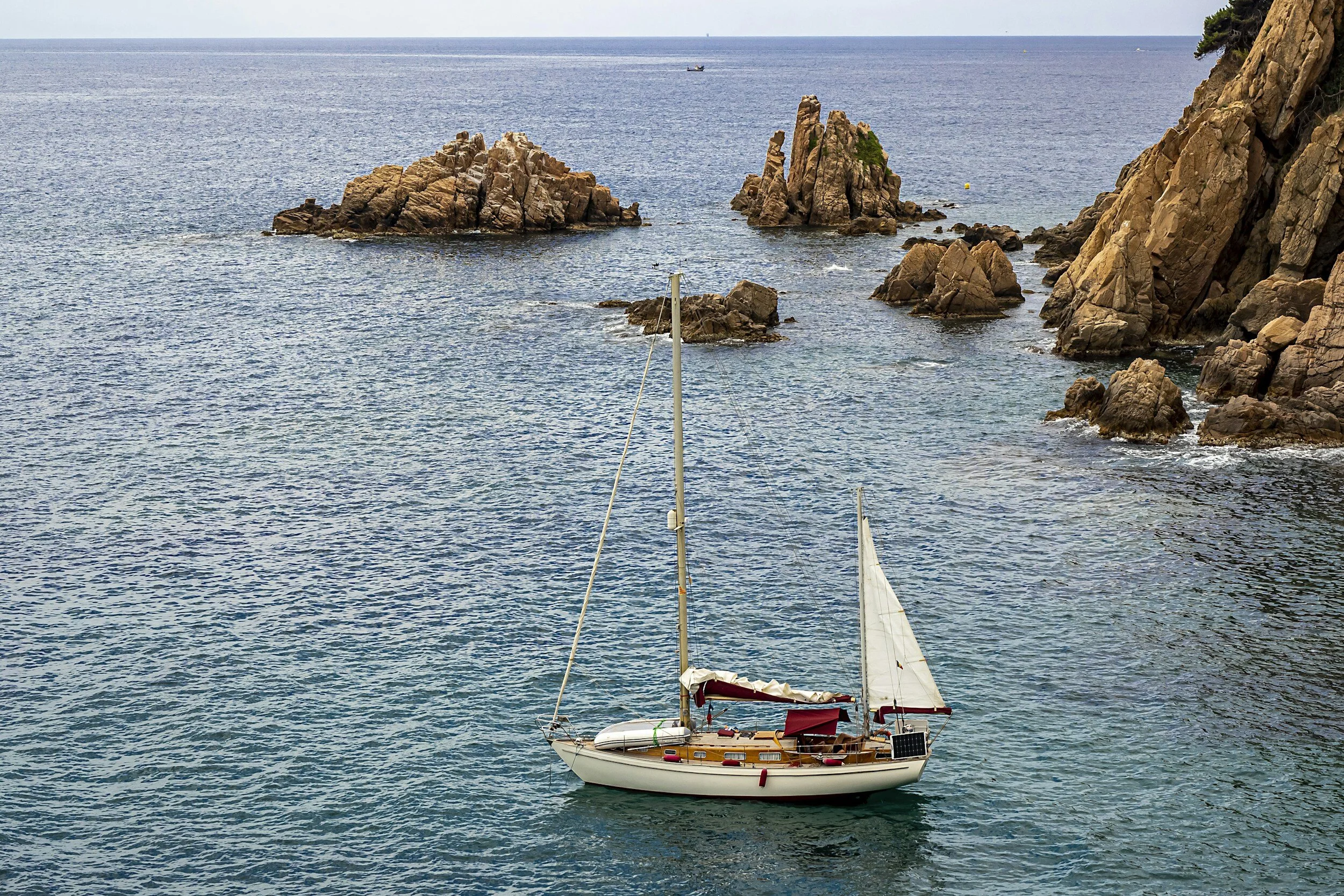 A sailboat near rocky cliffs on a calm ocean with several small rock formations in the water.