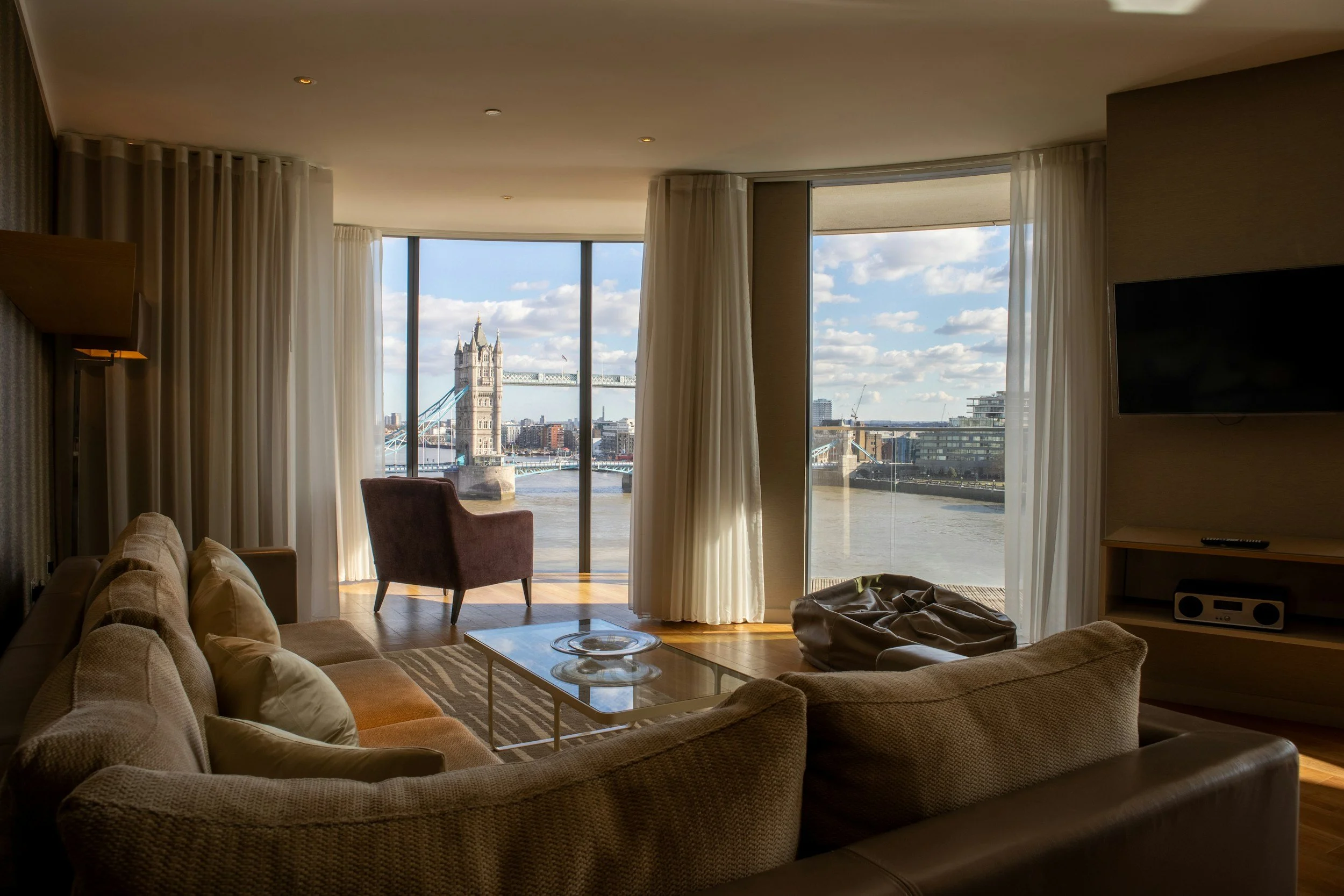 Living room with large windows showing the Tower Bridge and River Thames in London, beige sofa, pink armchair, glass coffee table, and flat-screen TV.