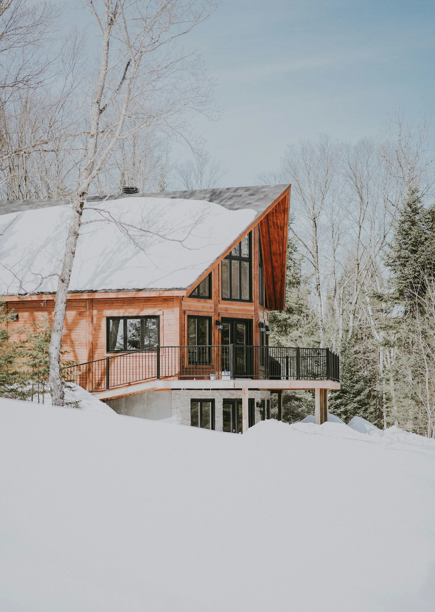 A modern wooden house with large windows and a balcony, surrounded by snow-covered ground and trees in winter.
