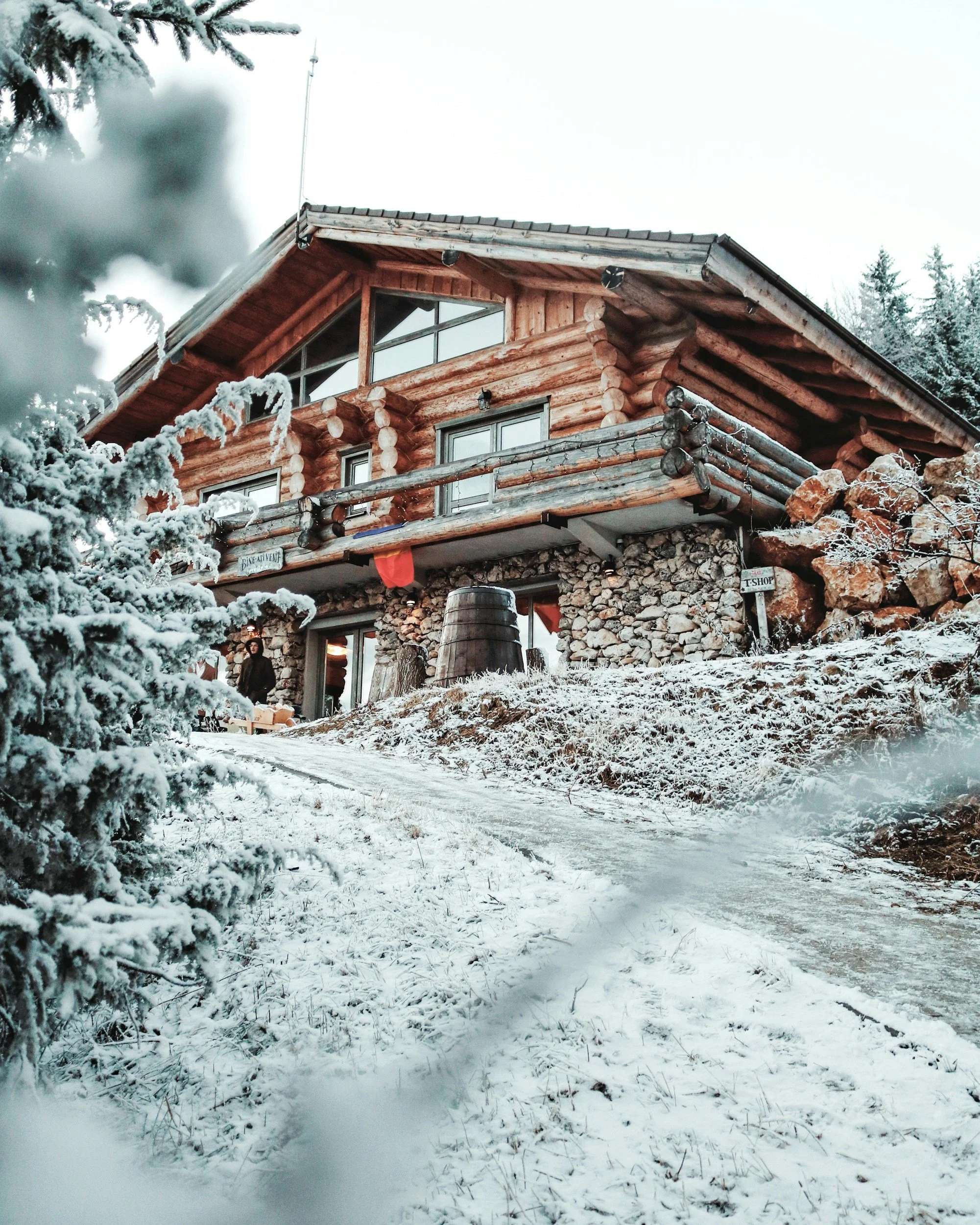 A two-story wooden cabin built with logs and stone, surrounded by snow, with a man standing on the porch.