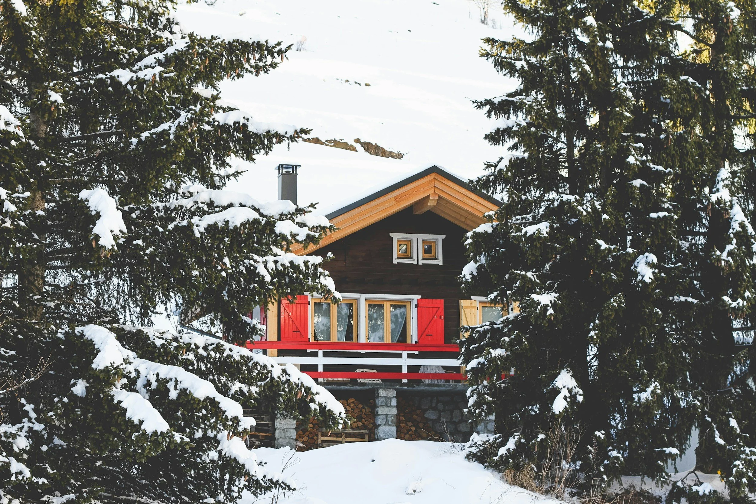 A cozy wooden cabin with a red and yellow door, surrounded by snow-covered pine trees in a winter landscape.