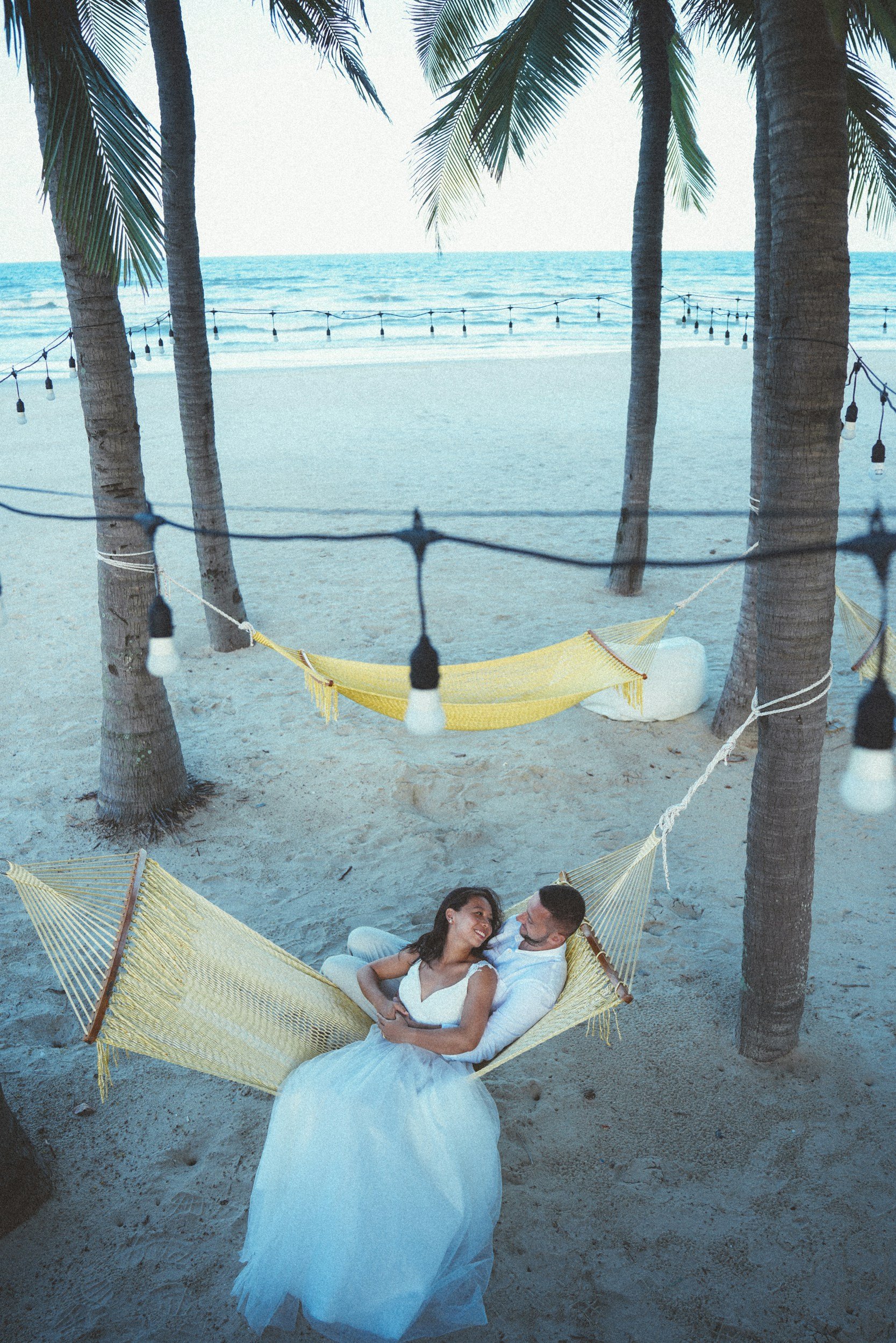 A couple lying in a hammock between palm trees on a sandy beach, with the ocean and string lights in the background.