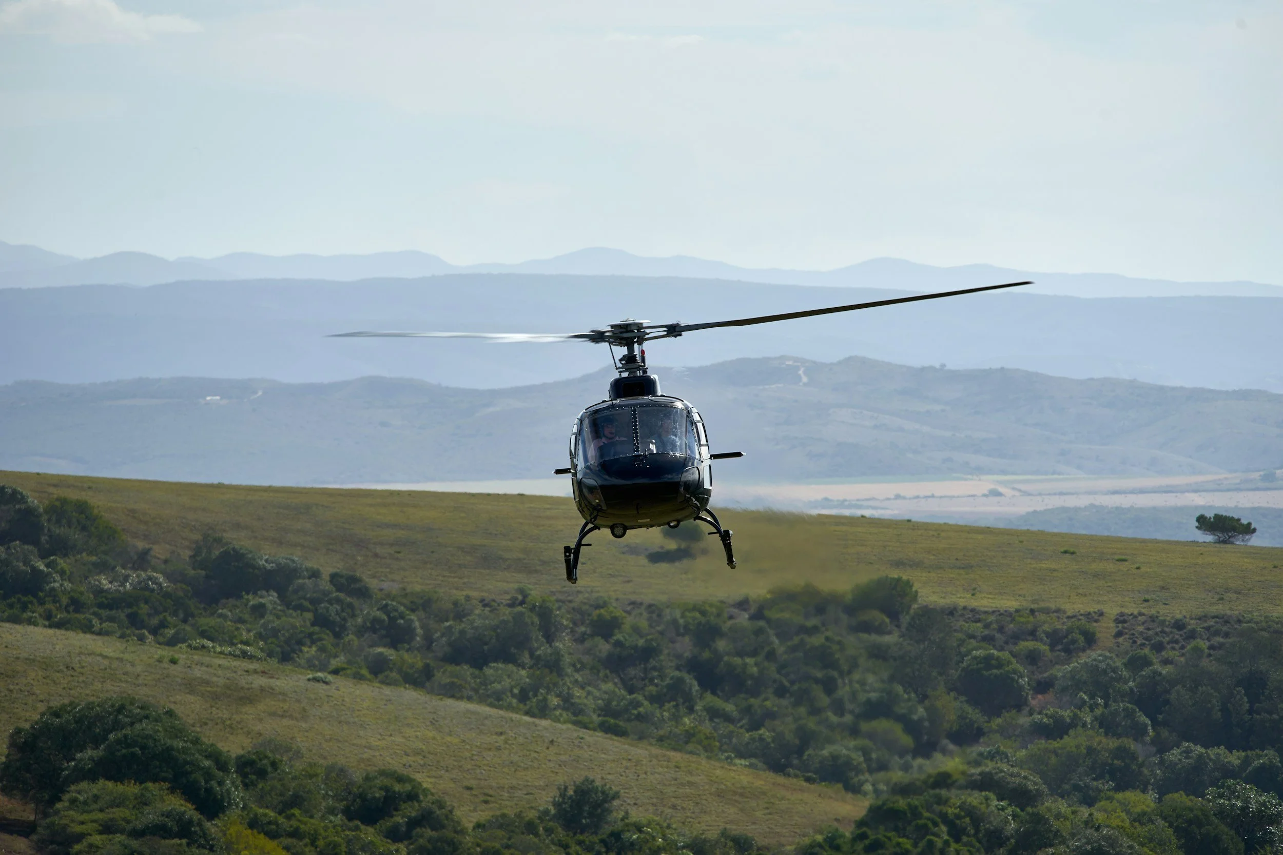 A black helicopter flying over a green landscape with hills and trees in the background.