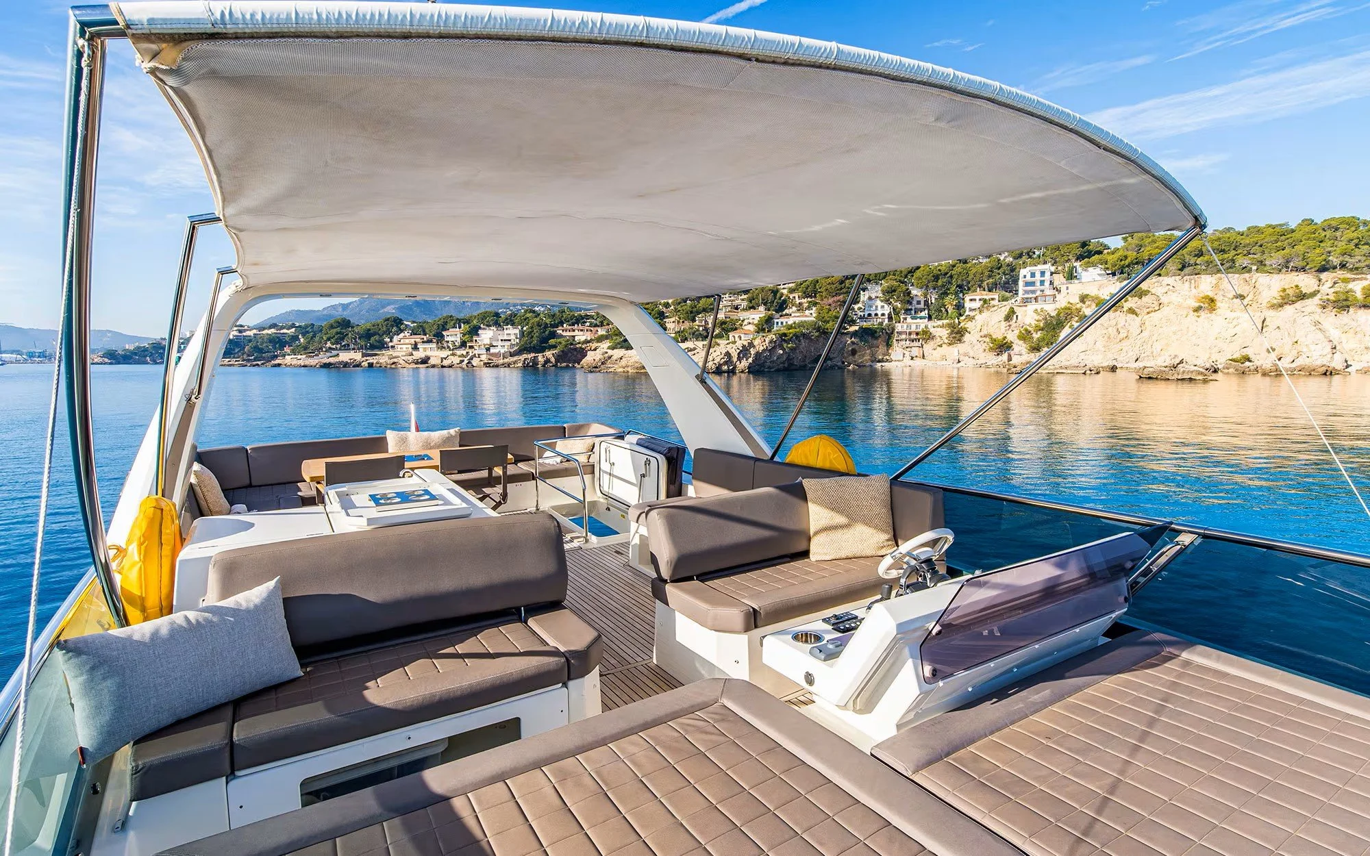 Luxury yacht deck with cushioned seating, a table, and a view of the coastline with houses and greenery.