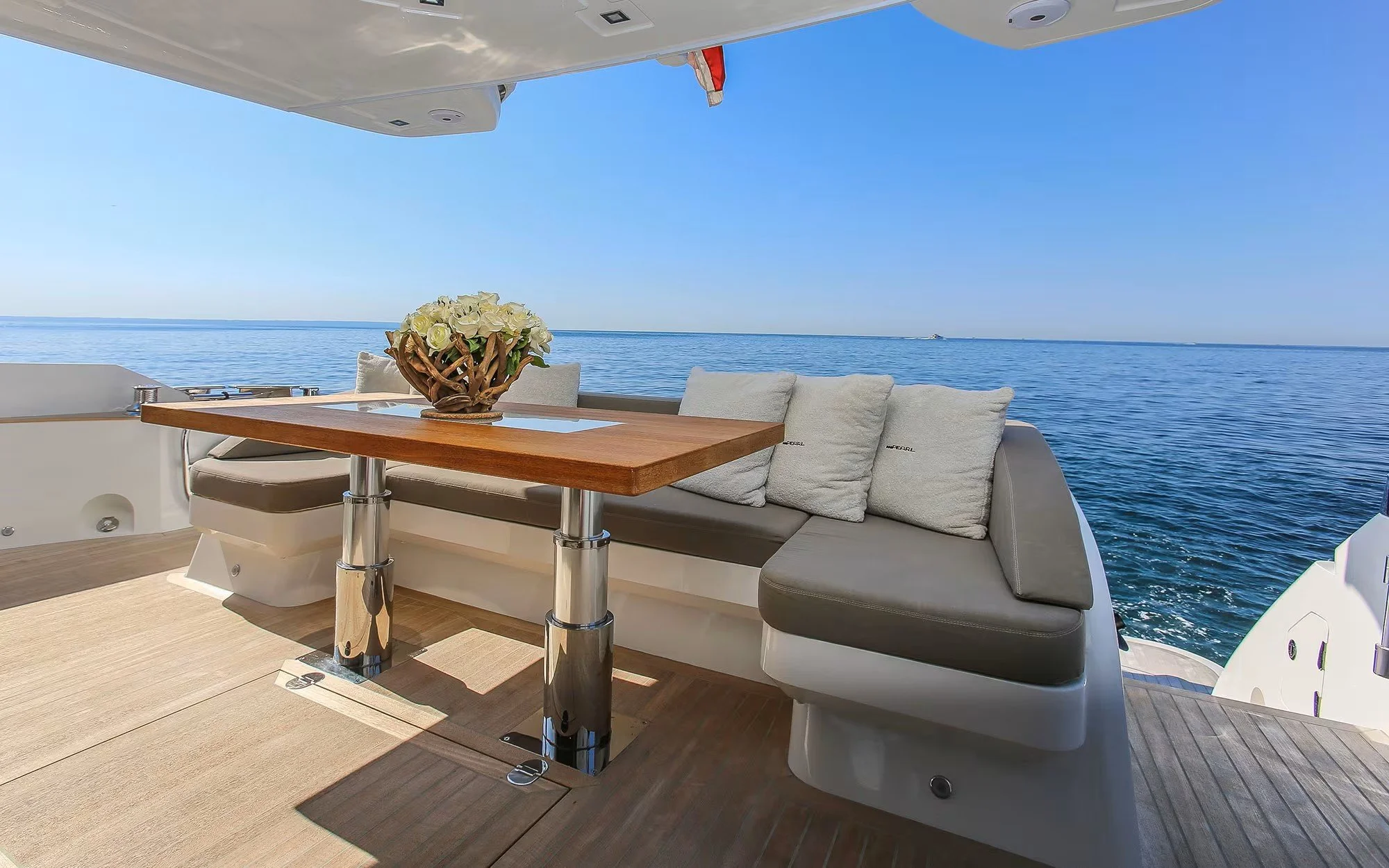View of a yacht deck with a wooden table, floral centerpiece, cushioned seating, and a view of the ocean under a clear blue sky.