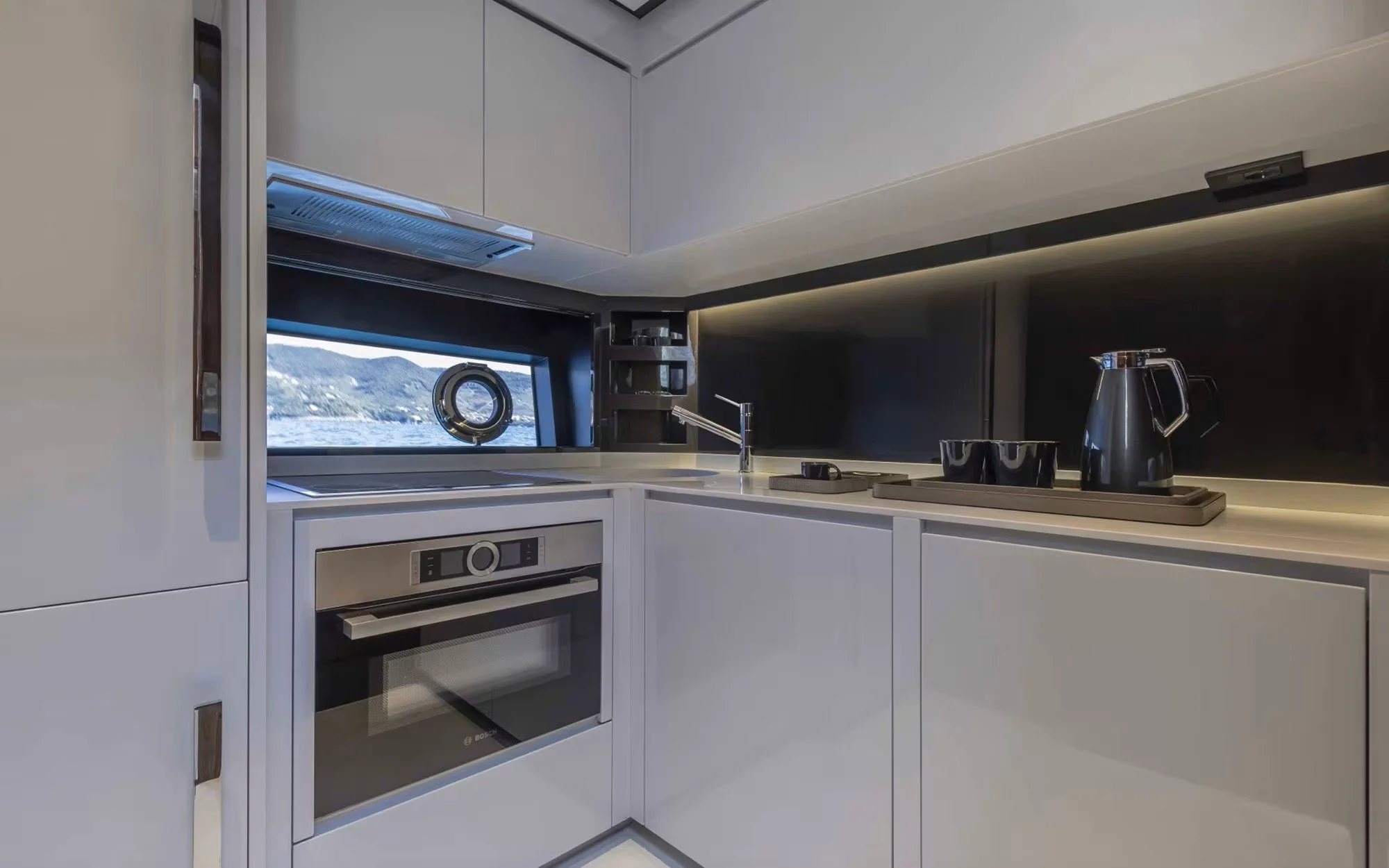 Modern compact kitchen with white cabinets, countertop, oven, black backsplash, sink, and window showing water and mountains outside.
