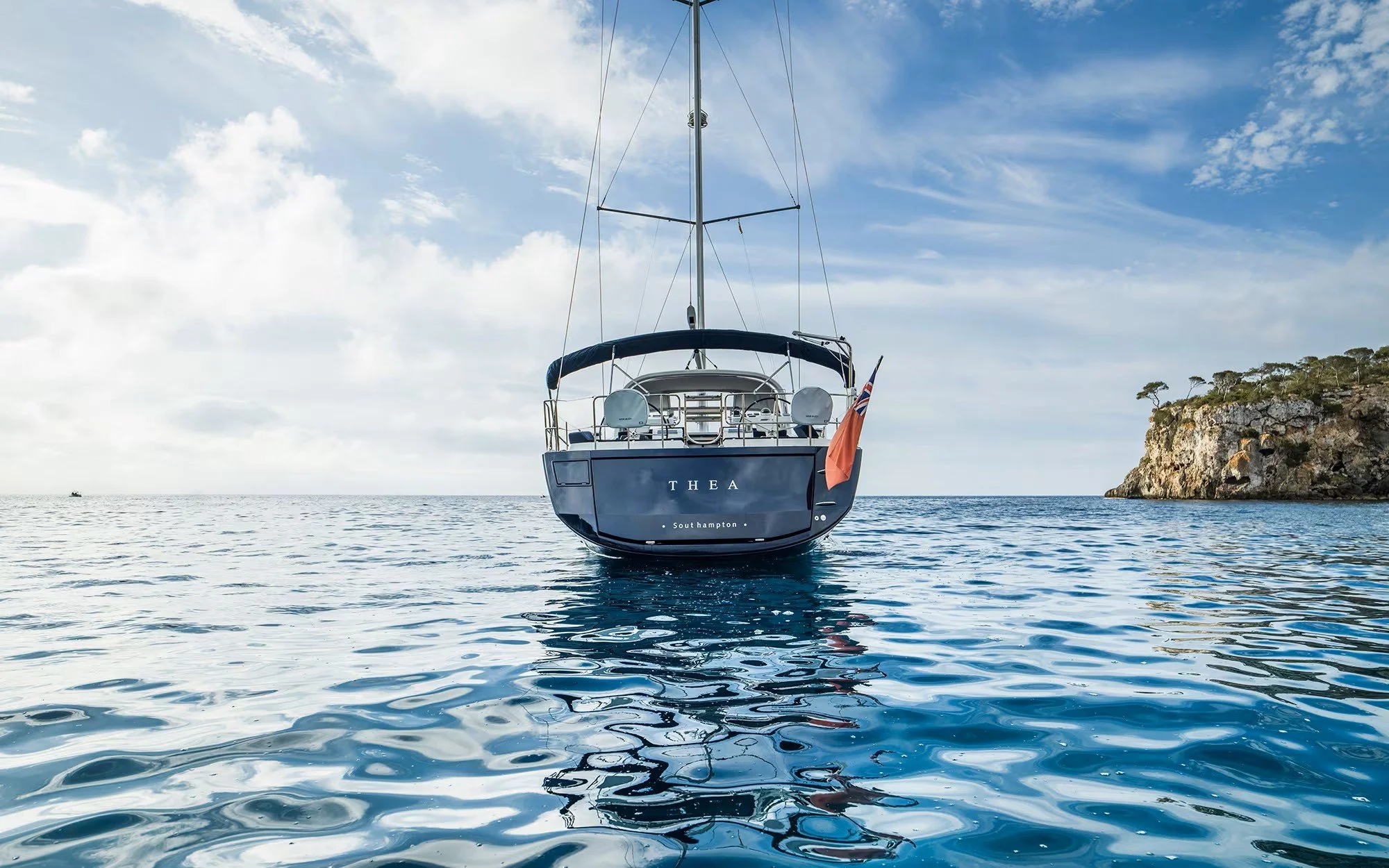 A sailboat named Thea floats on calm water near a rocky island with a clear sky in the background.