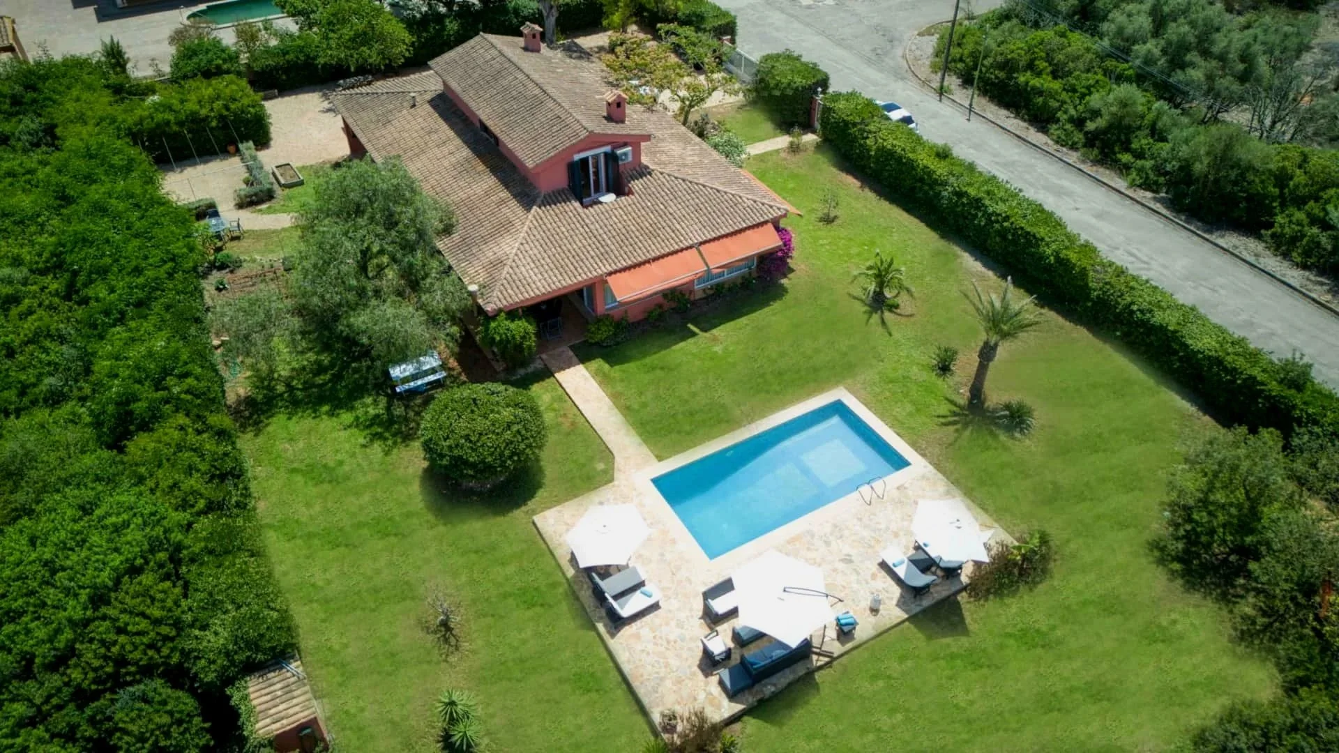 Aerial view of a house with a pool, surrounded by greenery and trees, with outdoor furniture and umbrellas near the pool.