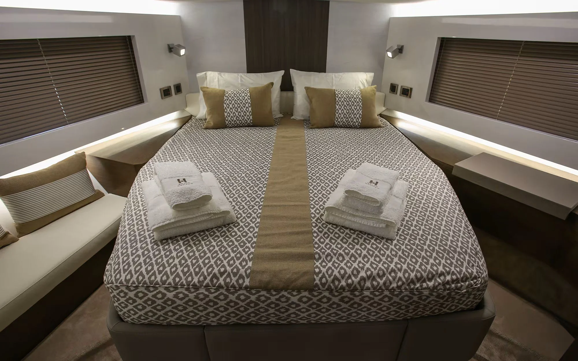 Hotel room with two twin beds pushed together, each topped with folded towels, pillows, and a patterned bedspread. There is a sofa with cushions on the left, and window blinds on either side of the beds. Small side tables and wall-mounted lamps are v