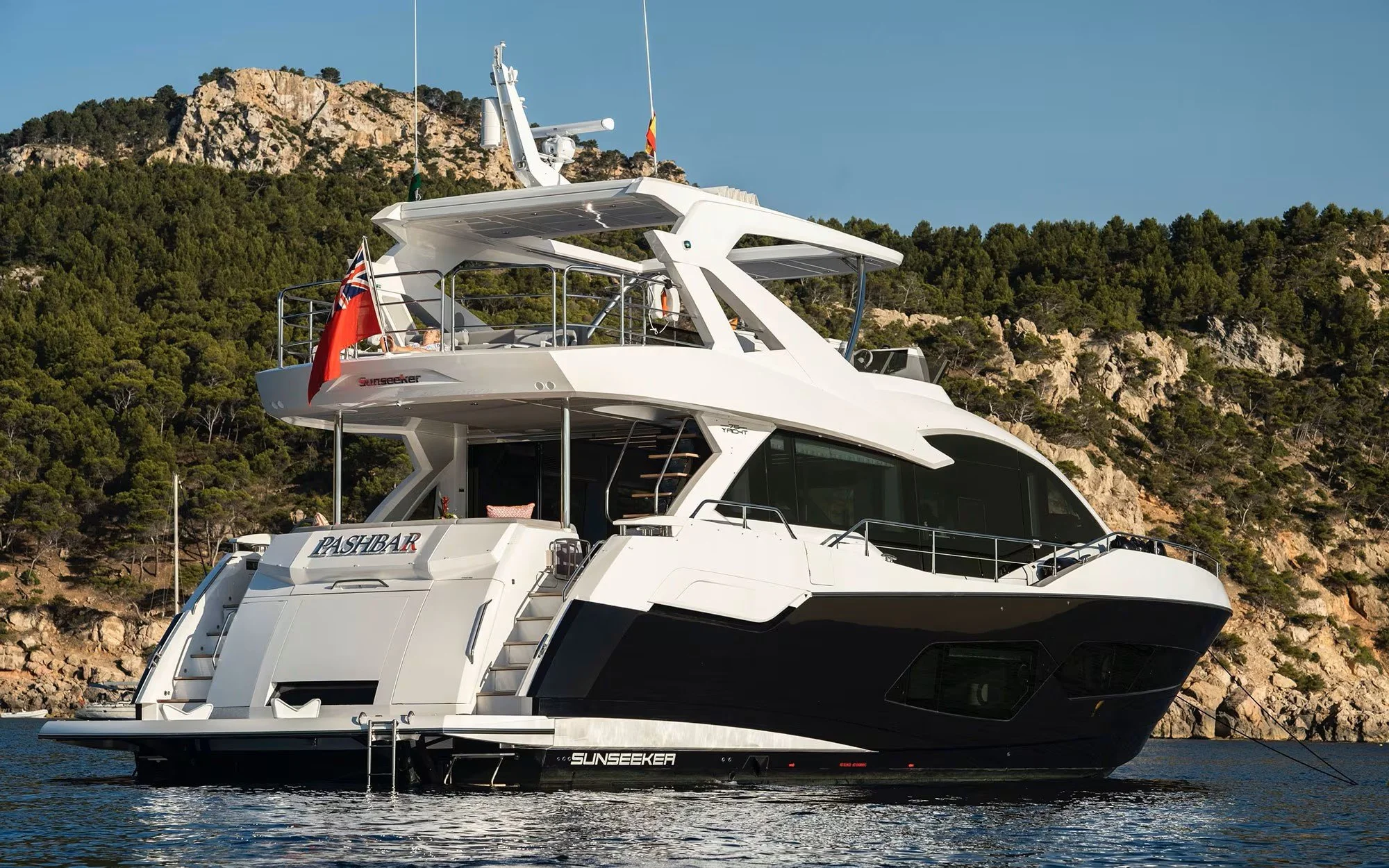 Modern white yacht named Sunseeker Pashbar floating on water with rocky hillside and trees in the background.