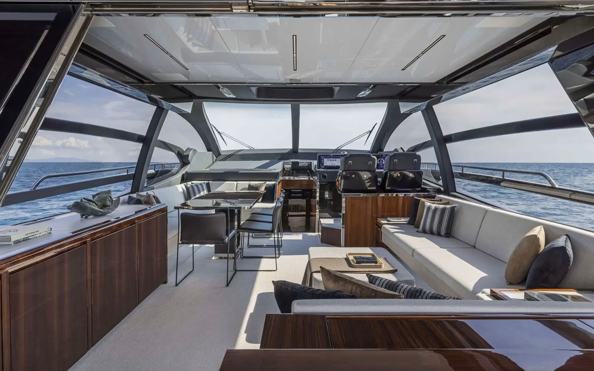 Interior of a luxurious yacht with a sitting area, dining table, and navigational controls, overlooking the ocean with open skies.