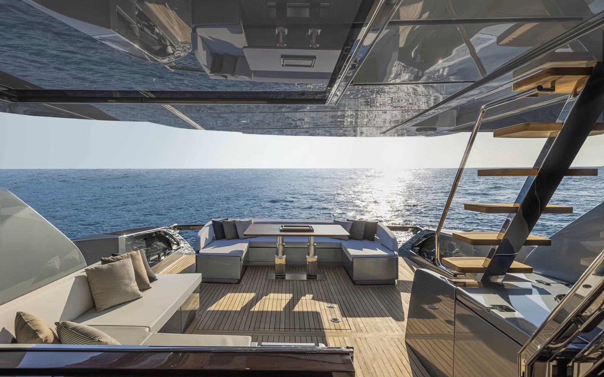 Luxury yacht's open deck with seating area, cushions, and stairs leading to an upper deck, overlooking the ocean with the sun in the background.