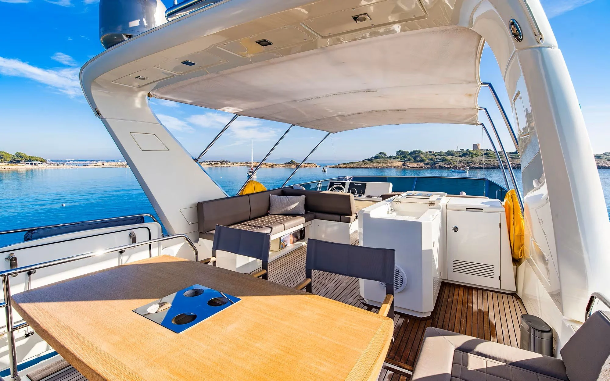 The deck of a yacht with seating and a dining table, overlooking calm waters and distant islands under a partly cloudy sky.