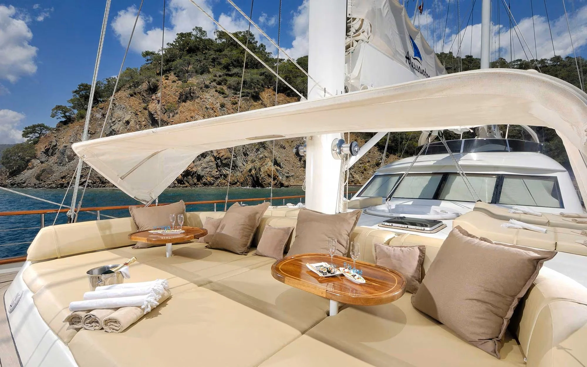 Luxury yacht with a shaded seating area featuring pillows and small tables with drinks, sailboat rigging, rocky shoreline, and blue sky in the background.